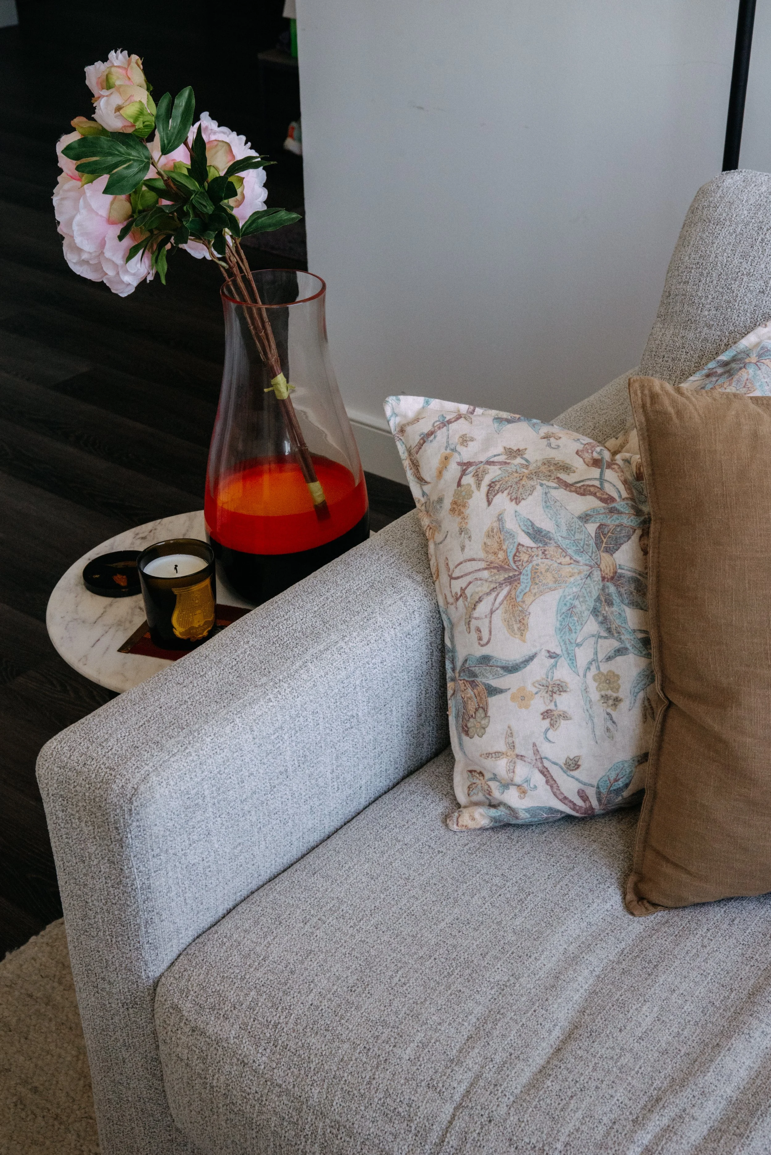 Living room corner with a light gray upholstered armrest, two decorative pillows, a marble side table with a candle and coaster, and a large glass vase with pink and white peonies on the table.