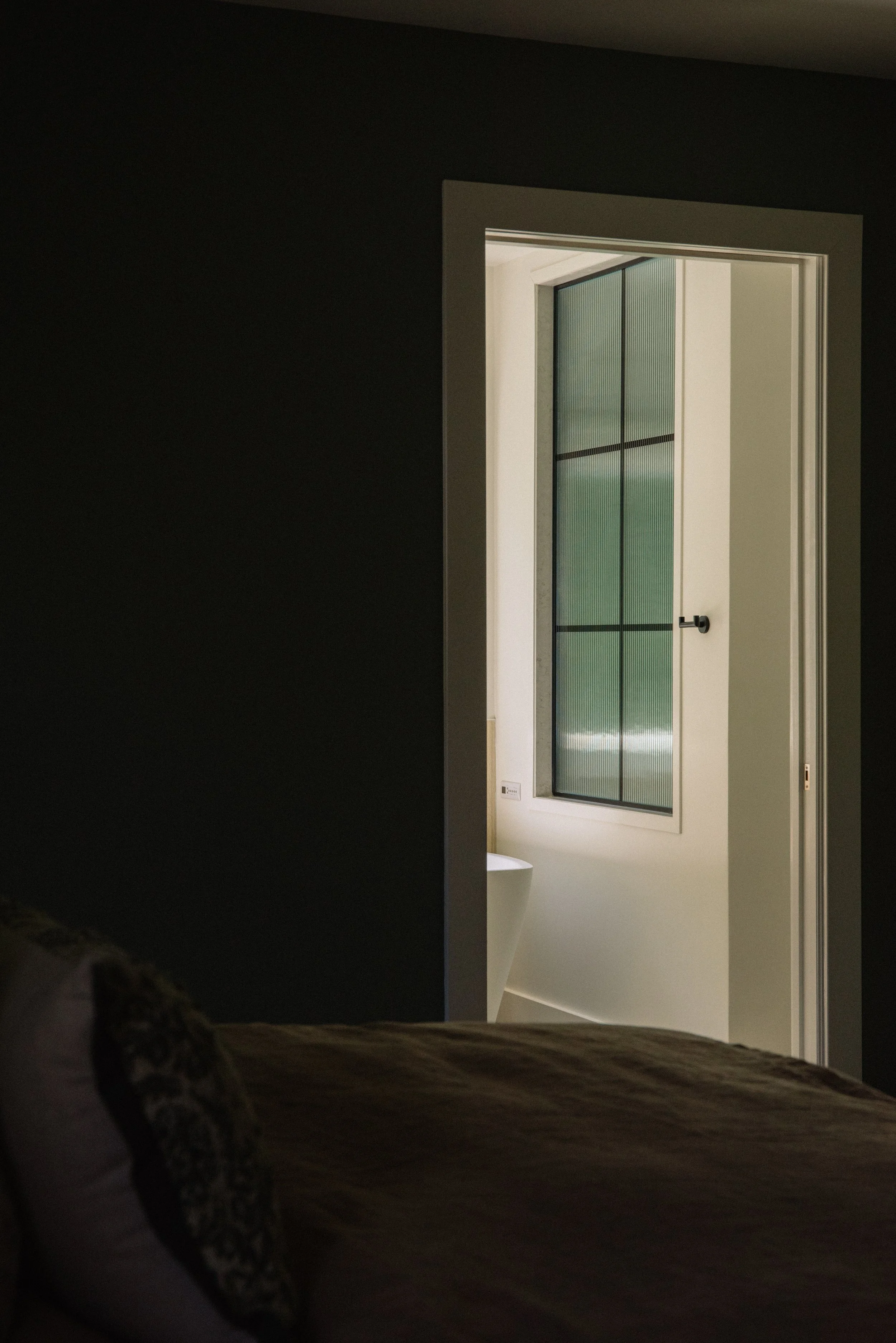 A bedroom with a dark wall, a bed with pillows, and a door leading to a bathroom with a window and a pedestal sink.