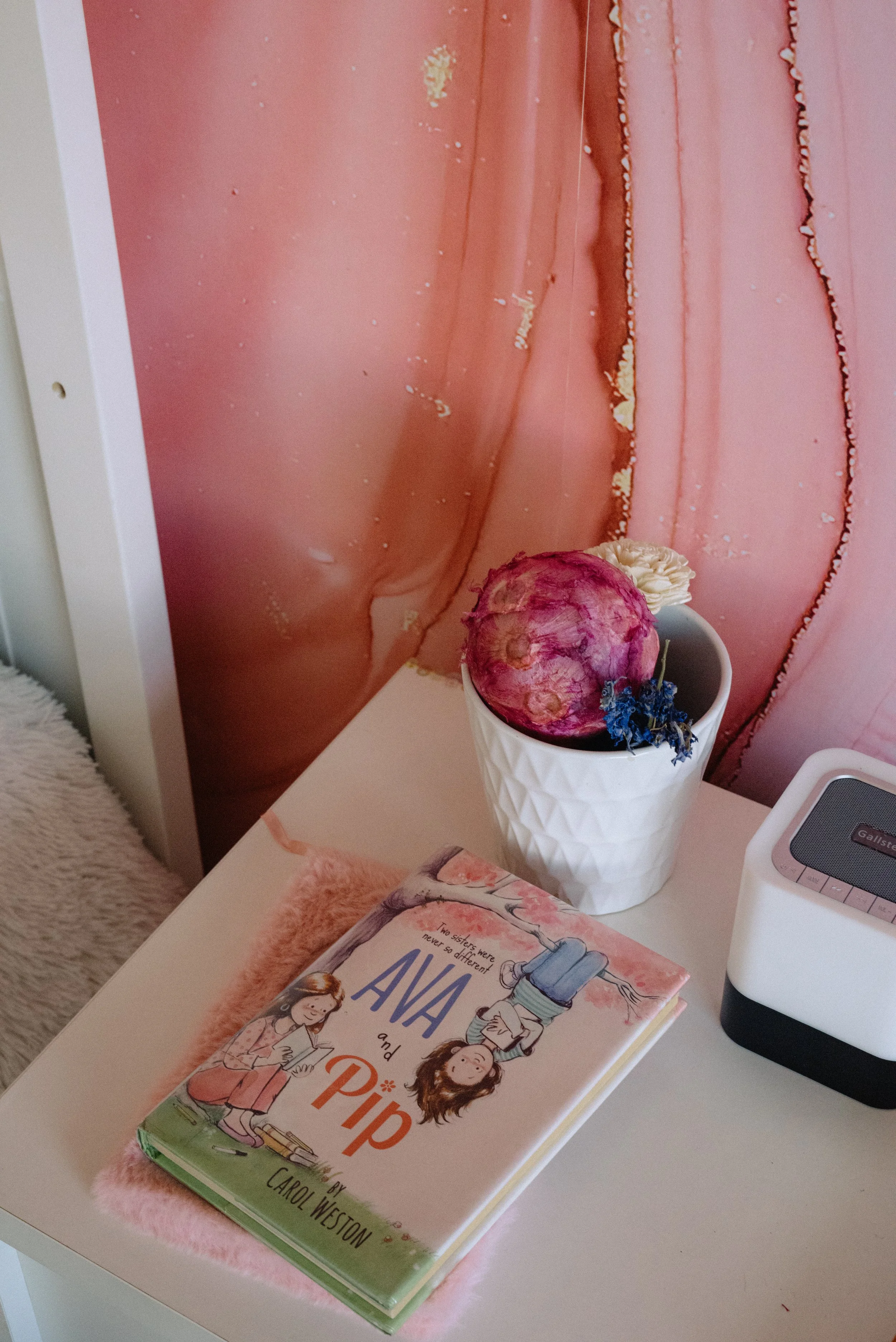 A white table with a pink book titled "Ava and Pip: Two Sisters Were Never a Difference" by Carol Weston, a white vase with a pinkish-purple ball of yarn and a white flower, and a small white speaker, set against a pink marble-patterned curtain.