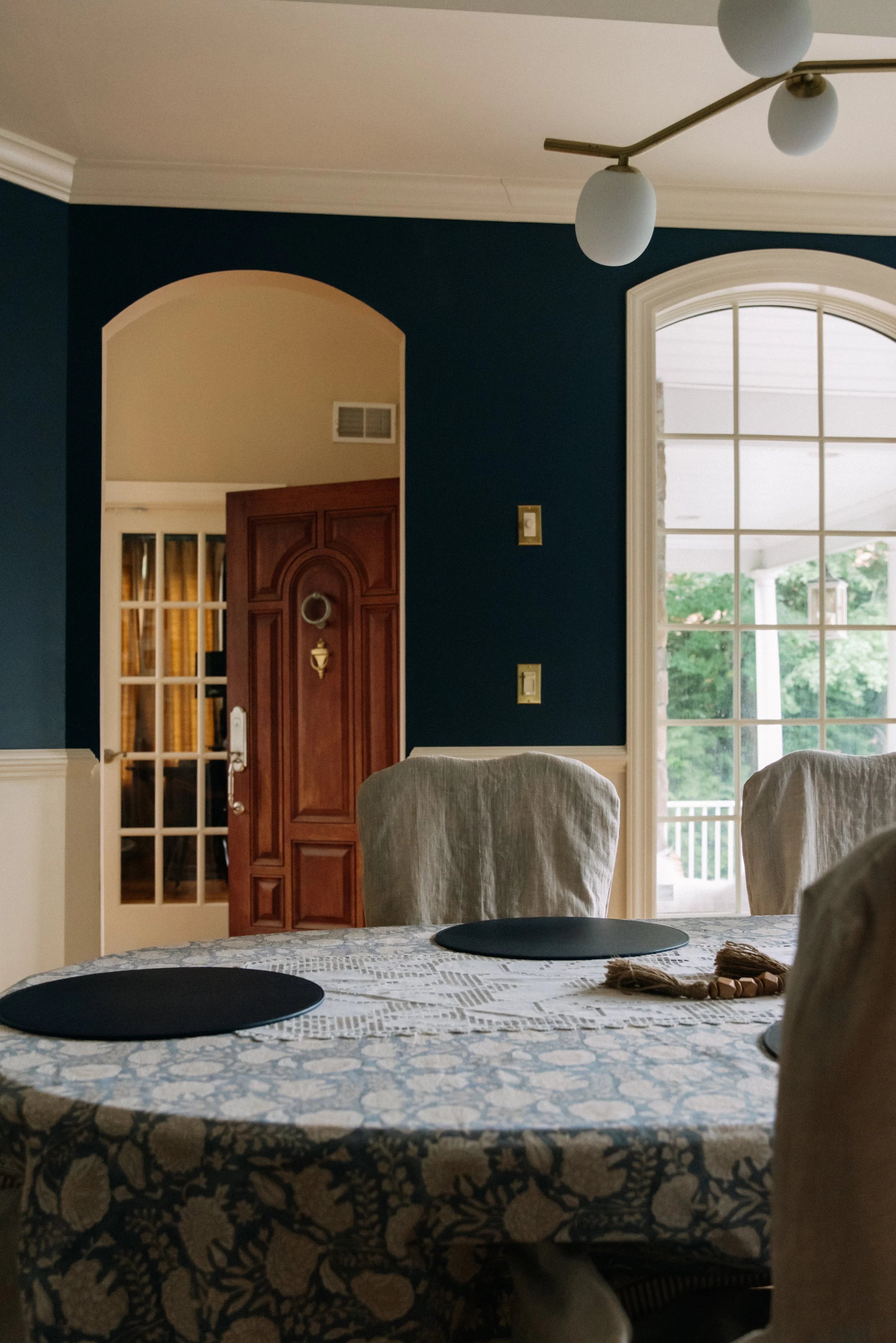A dining room with a round table covered with a patterned tablecloth, surrounded by chairs with cream-colored slipcovers. There is a large window with white framing letting in natural light, and a wooden front door with a circular window and a brass