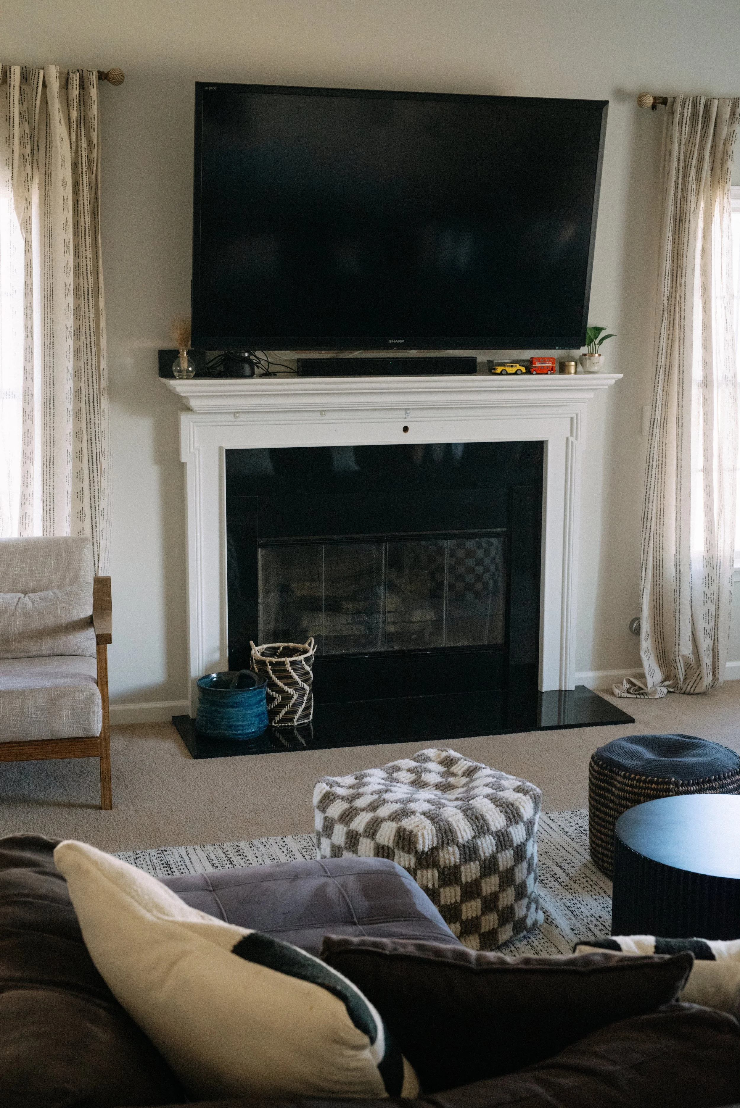 Living room with a white fireplace, flat-screen TV on top, decorated with small toys and a plant, beige curtains, and a sofa with pillows.