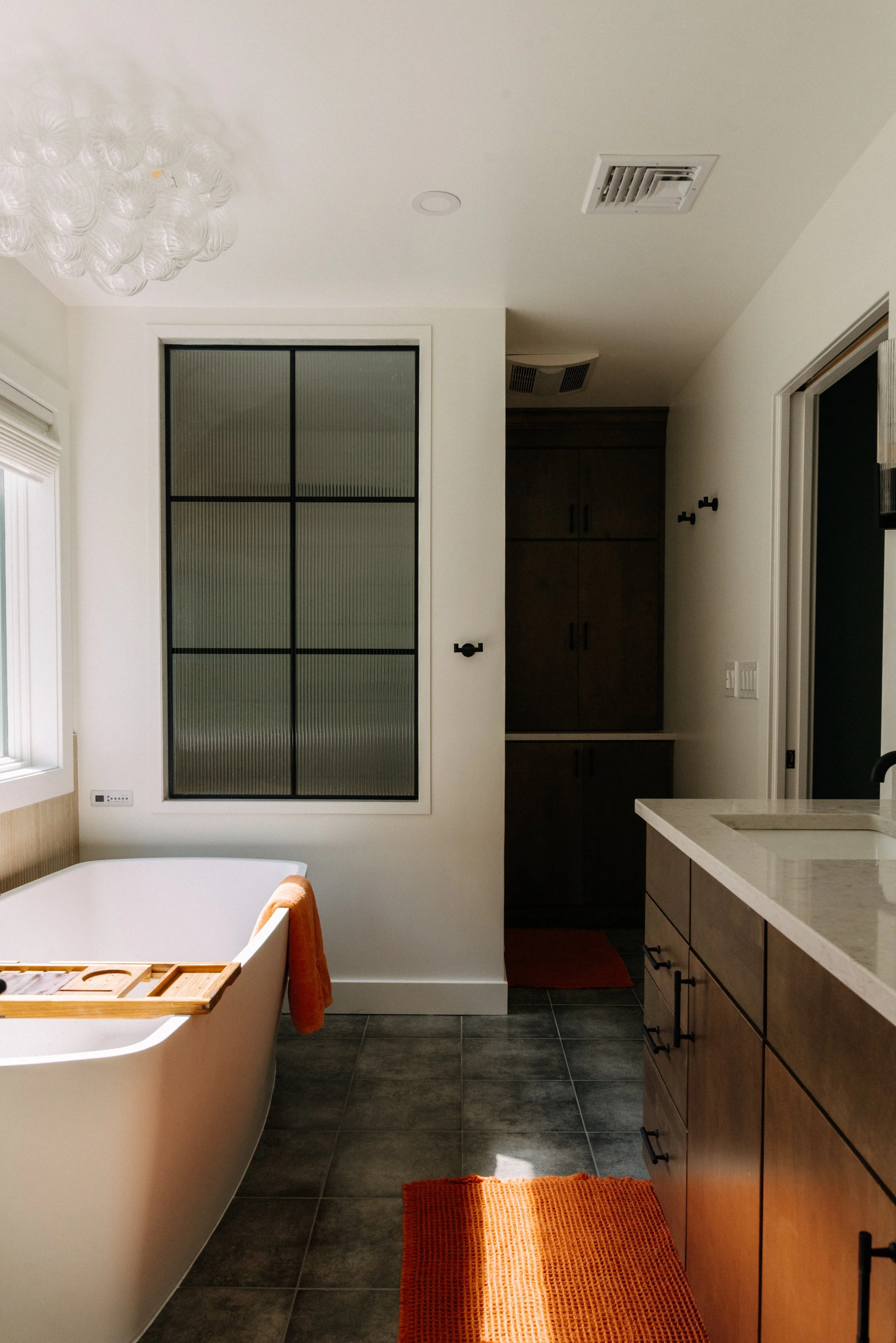 A bathroom with a white freestanding bathtub, a large window, dark gray floor tiles, and a wooden vanity with a white countertop. An orange towel hangs on the side of the bathtub, and an orange rug is on the floor.