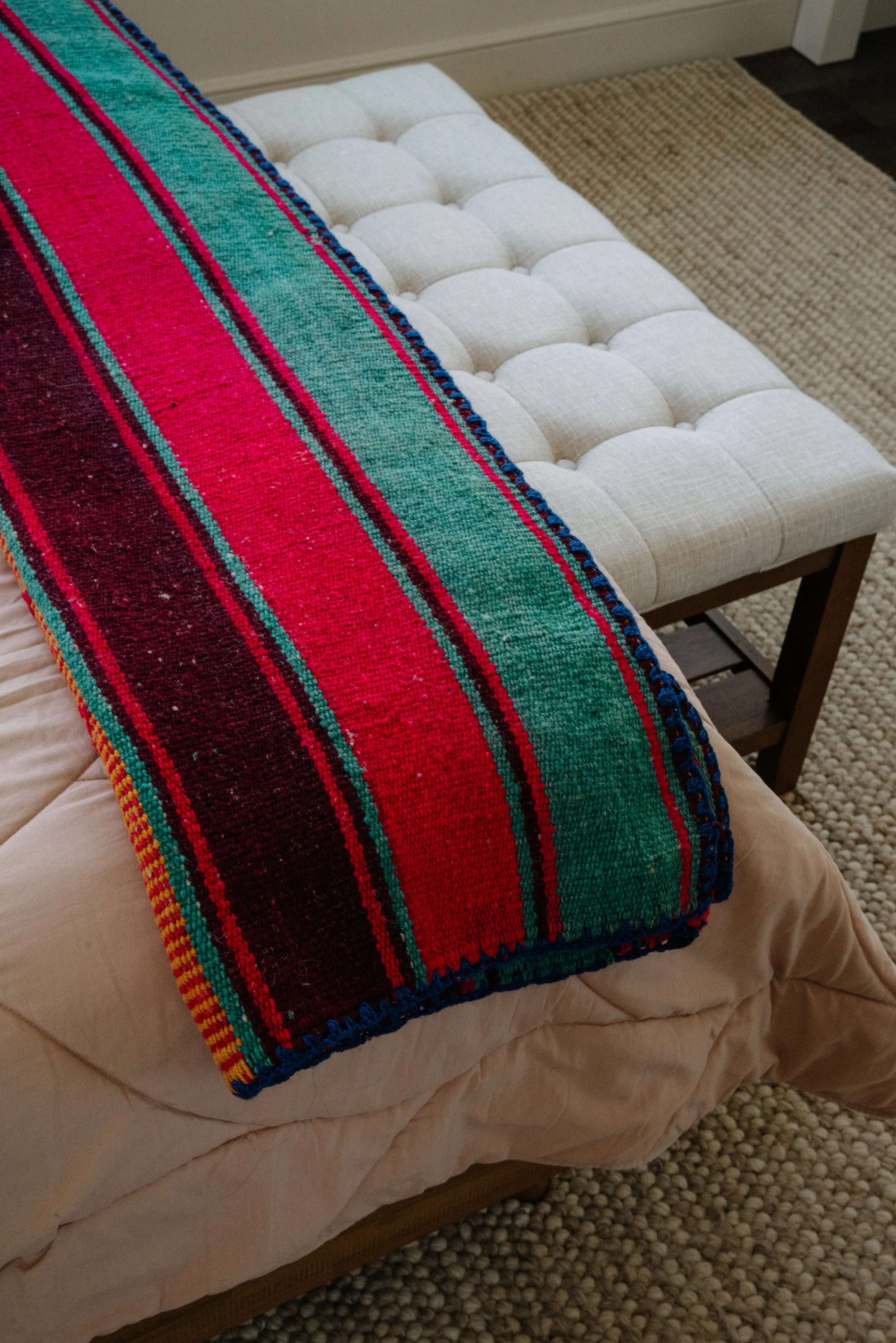 Colorful striped blanket draped over a cream-colored bed, with a white tufted ottoman and beige rug visible in the background.