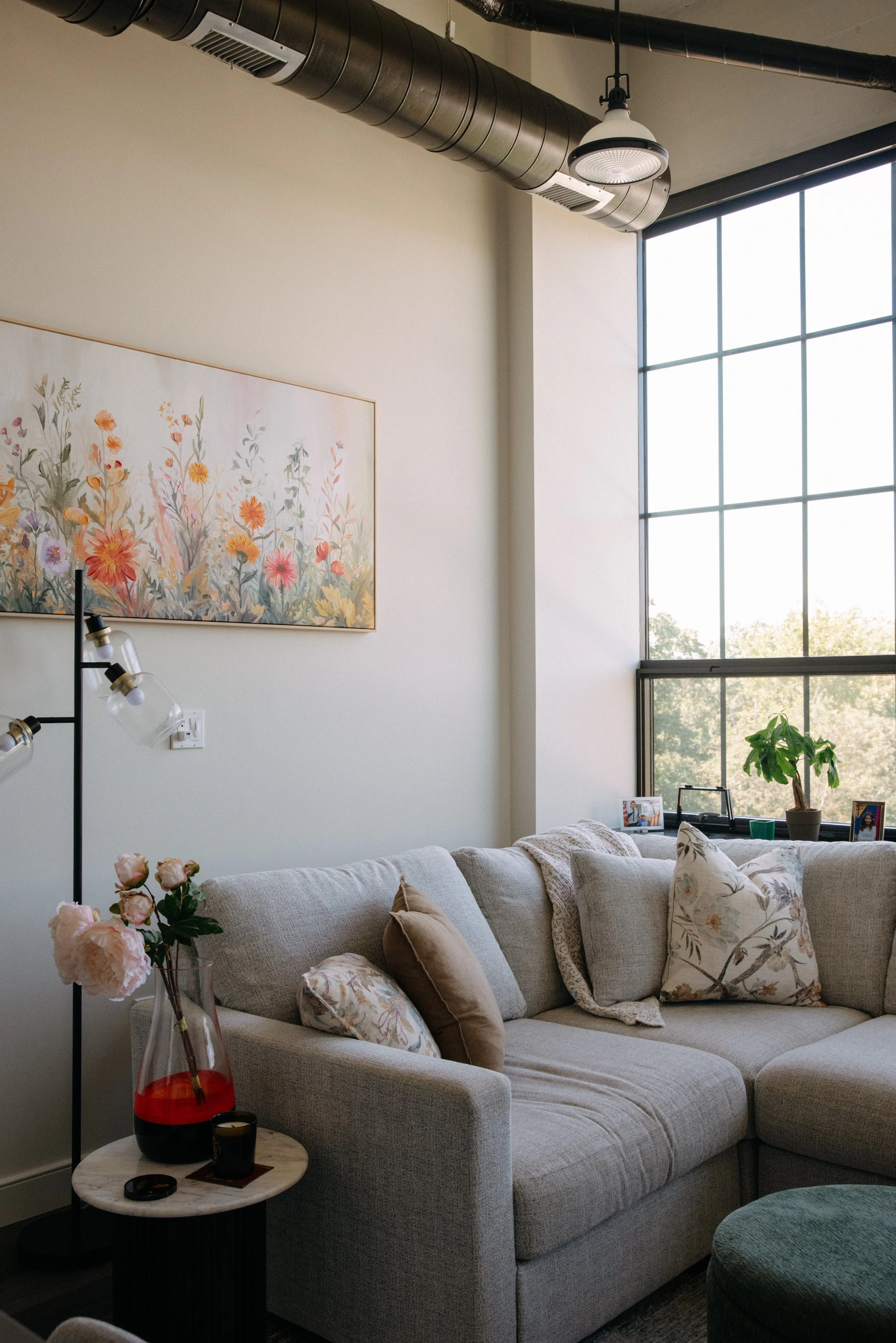 Living room with gray sectional sofa, decorative pillows, a small side table with a flower vase, framed photos, large window, floral artwork on wall, and industrial ceiling ductwork.