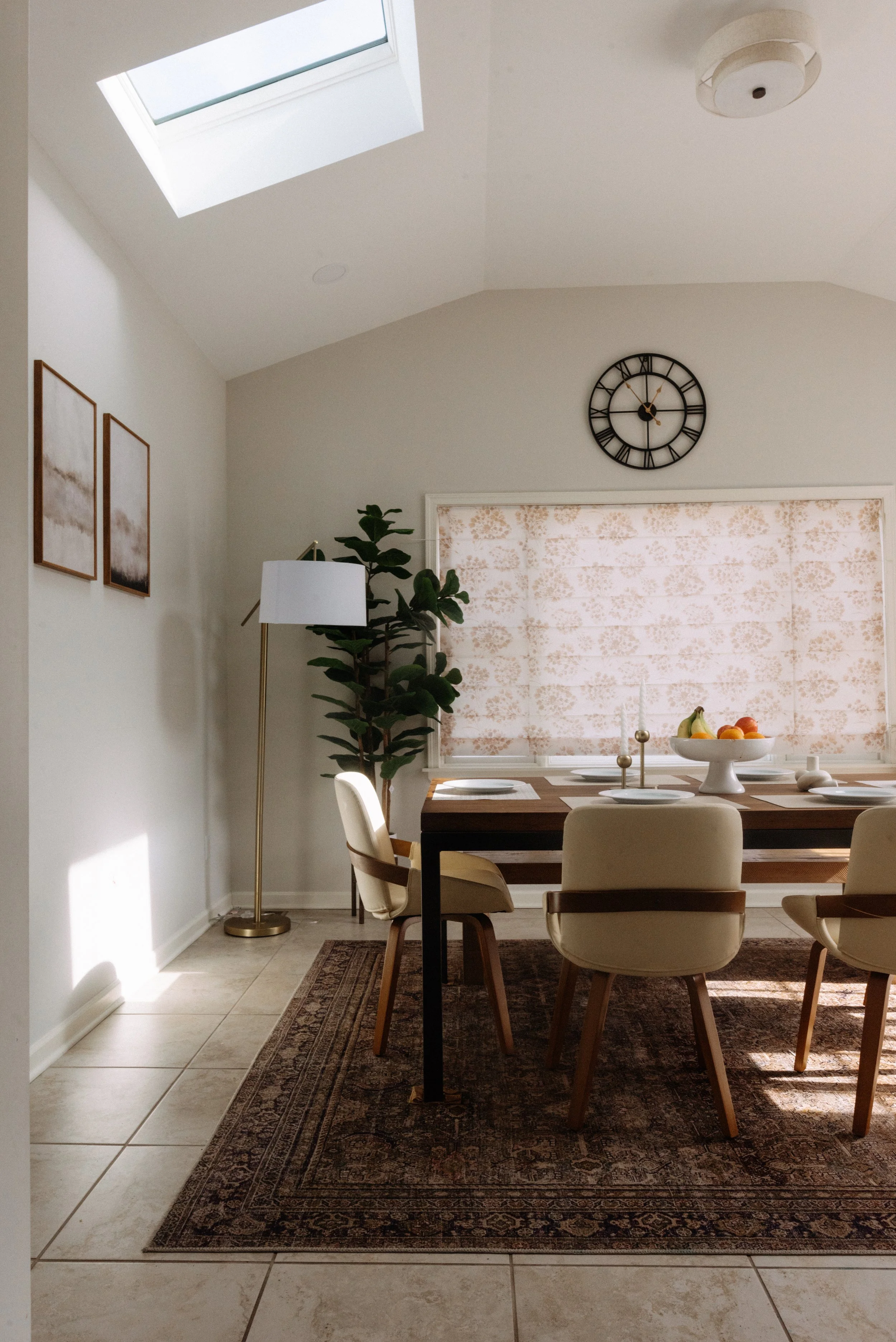 Dining room with beige chairs, a wooden dining table, a fruit bowl, wall art, a tall floor lamp, and a window with floral curtains, illuminated by natural light.