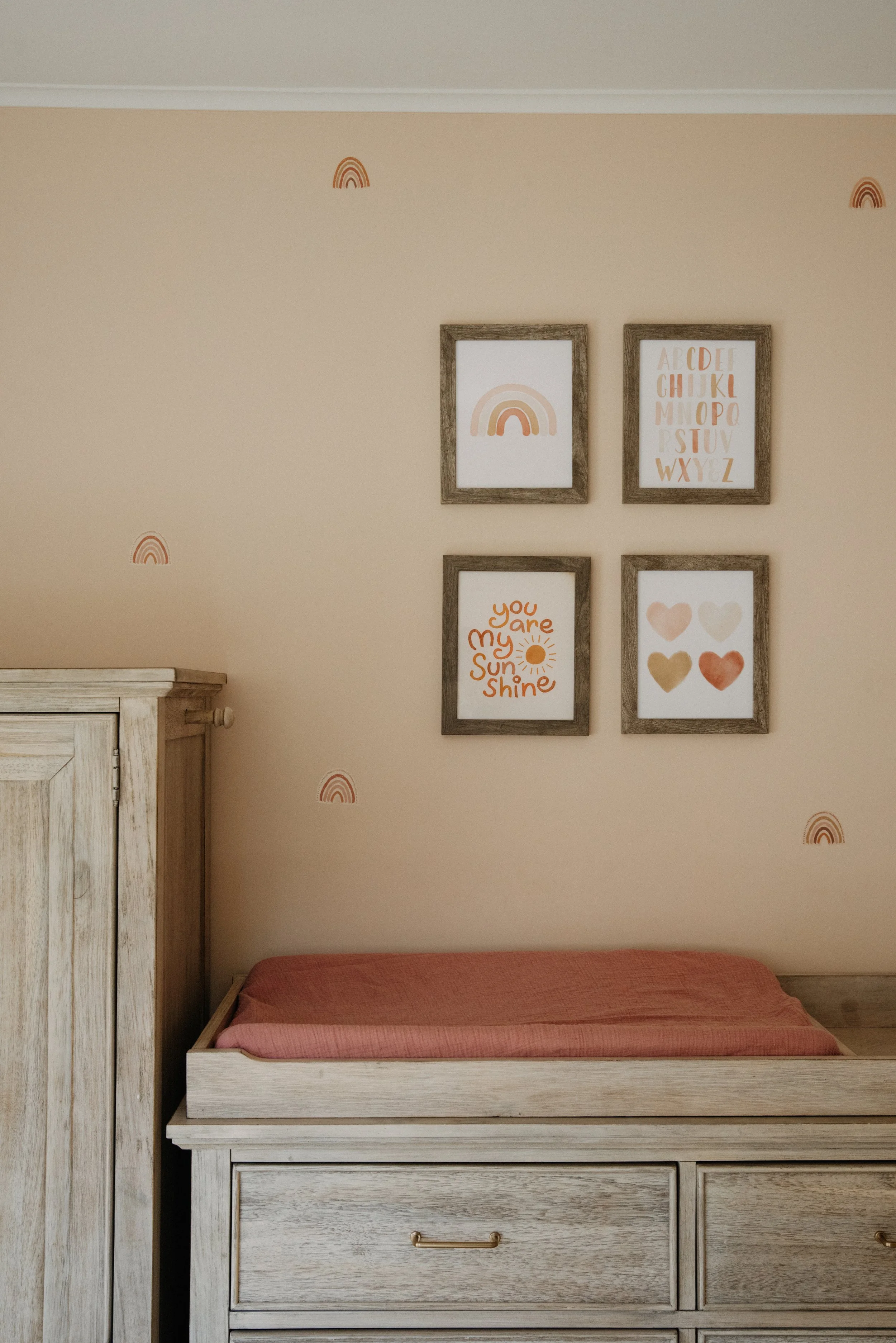 A wooden changing table with a pink changing pad, next to a matching wooden wardrobe, against a beige wall decorated with small rainbow decals. Above, four framed art prints with pastel colors and inspirational phrases or shapes.