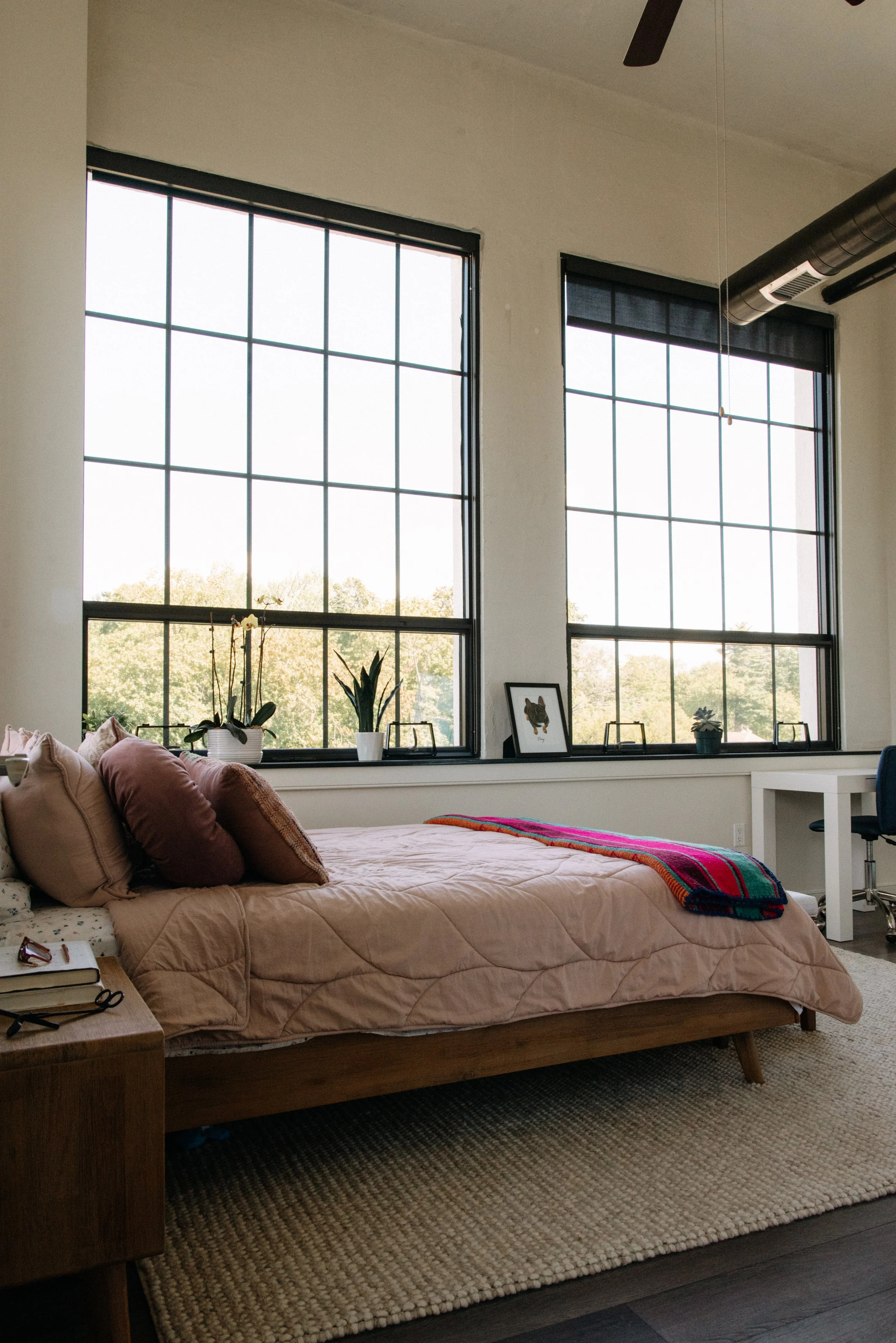A bedroom with a bed, pillows, nightstand, and large windows showing trees outside.