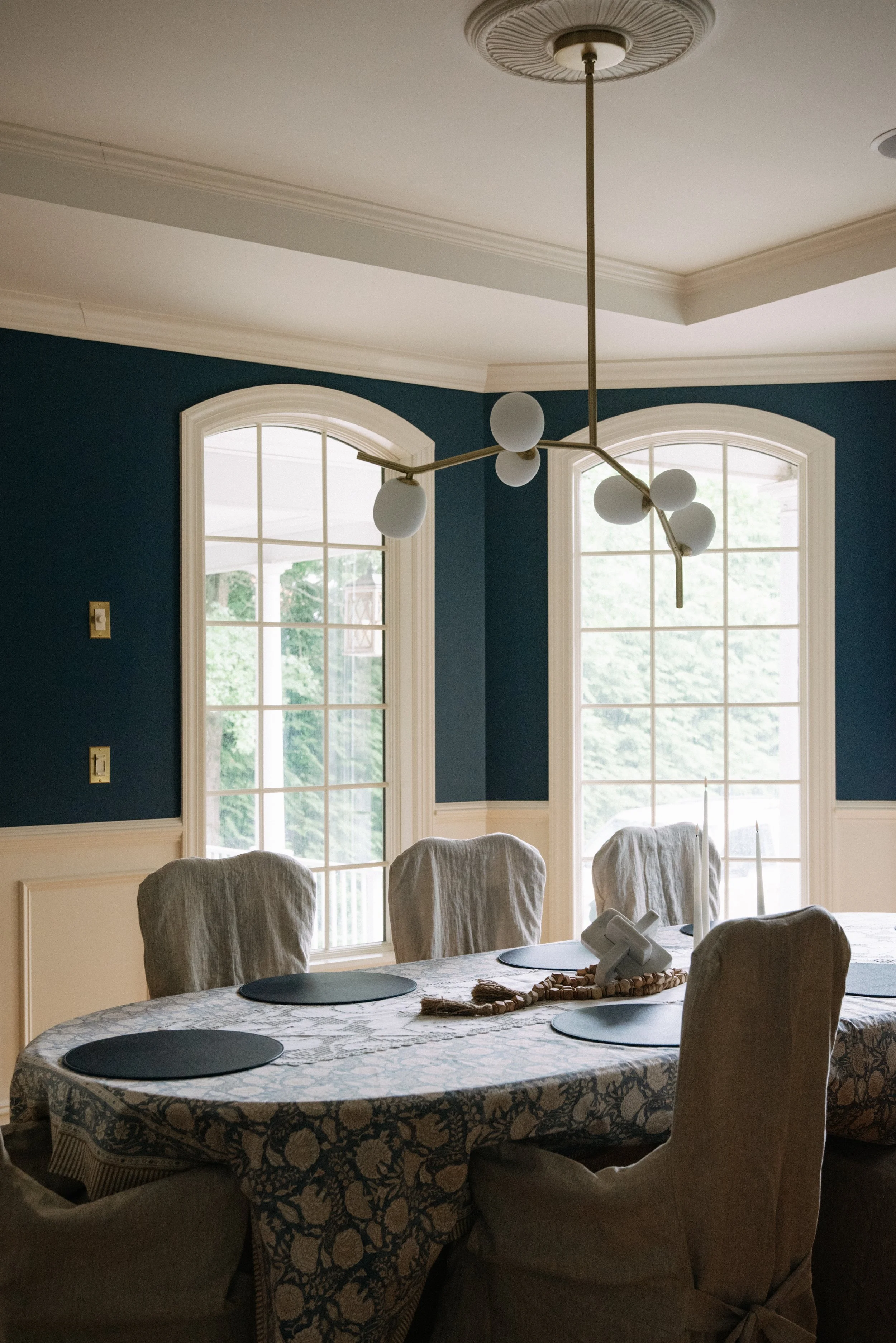 A dining room with a large oval table covered by a patterned tablecloth, surrounded by beige upholstered chairs. There are four black placemats on the table, and a decorative centerpiece with a telephone and candles. Behind the table, two large windo