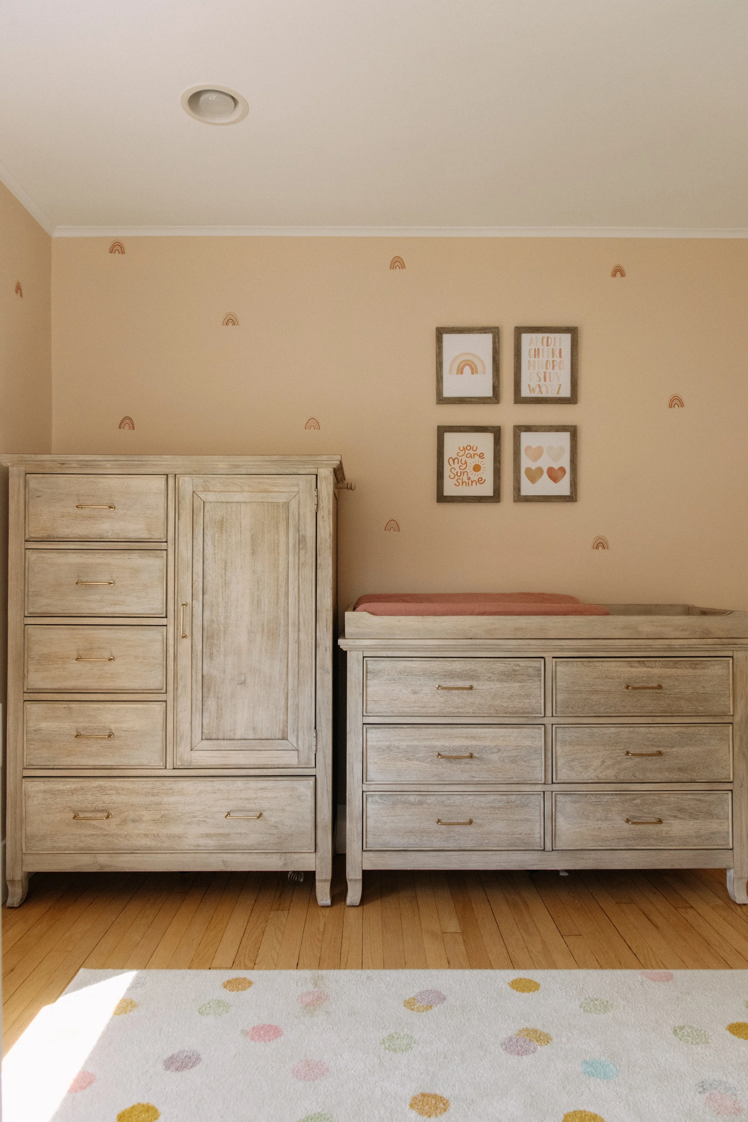 A nursery with a wooden dresser and changing table, wall art with pastel colors, and a white rug with colorful polka dots.