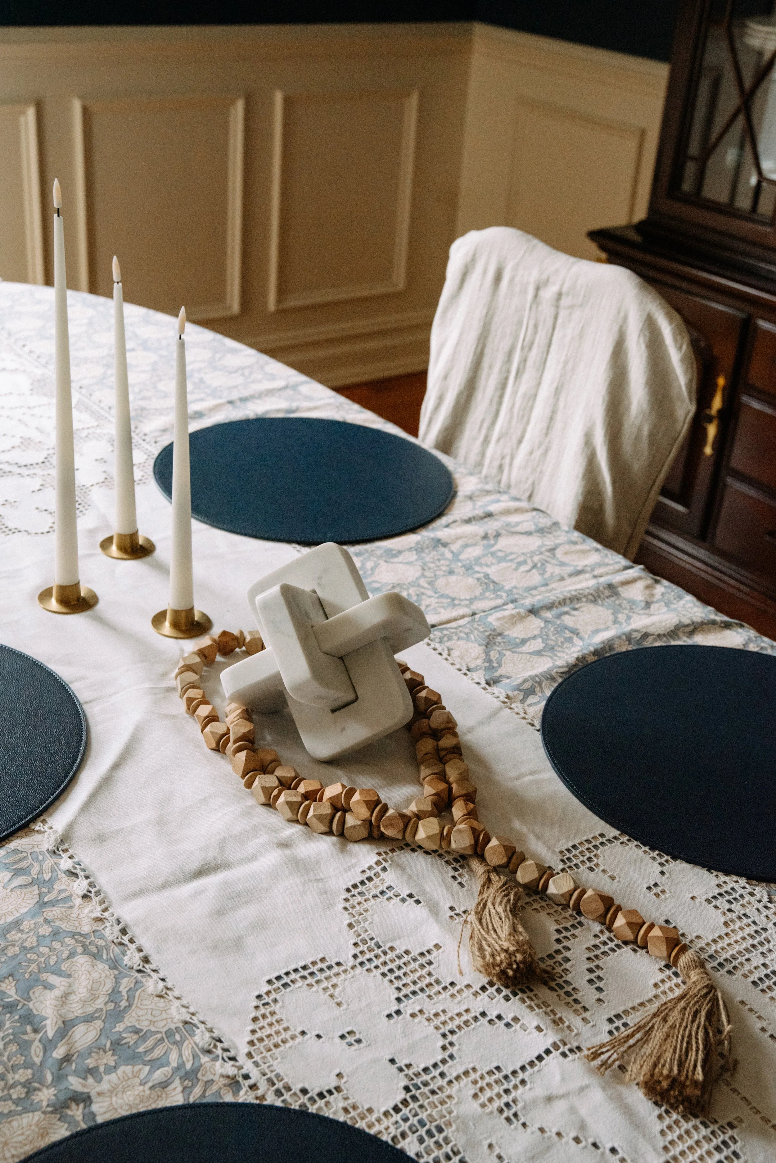 A dining table with a lace tablecloth, three navy blue placemats, three white candles in brass holders, a white ceramic sculpture, and a wooden beaded necklace. A cream-colored upholstered chair is seen in the background.