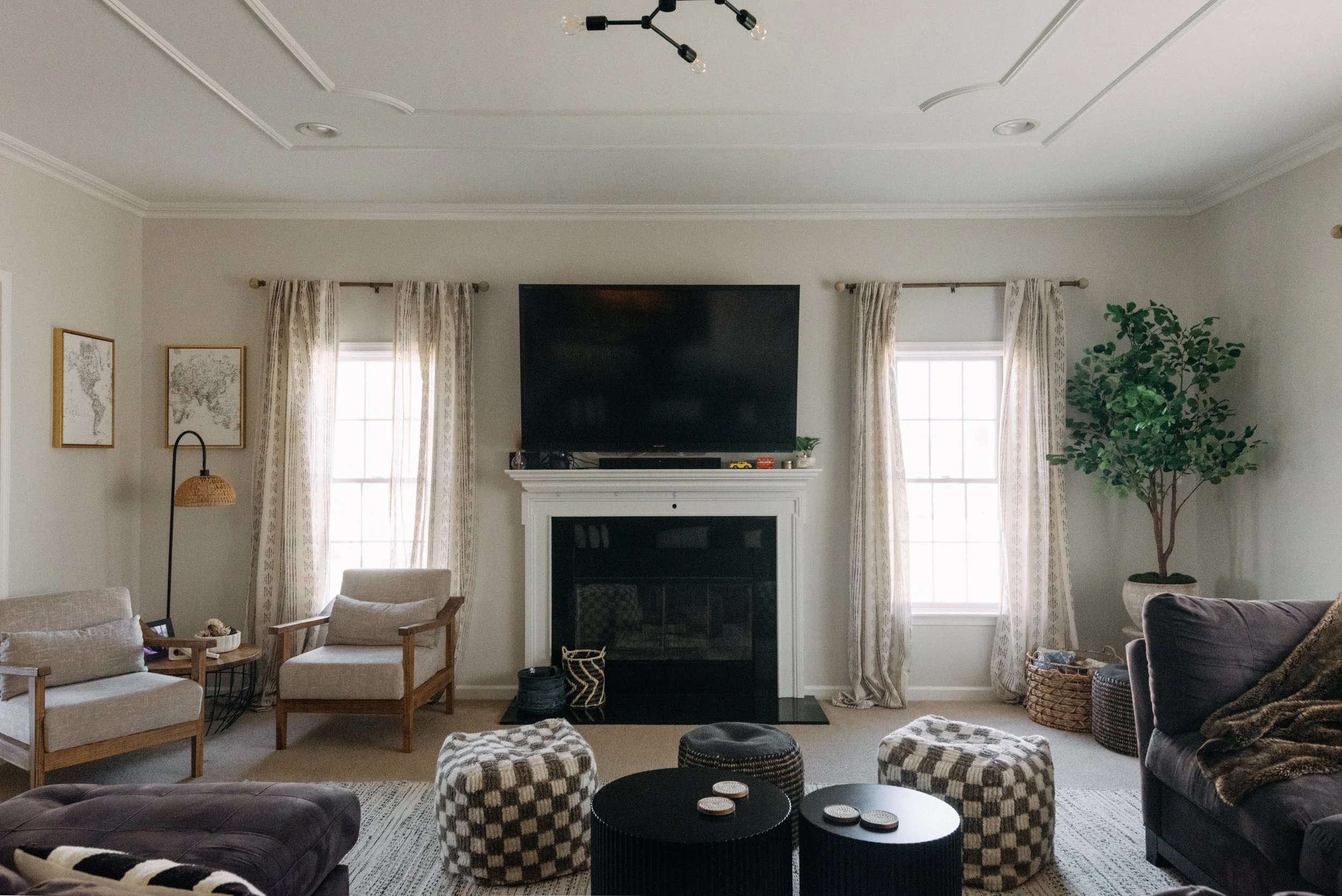 Living room with beige walls, white crown molding, two windows with patterned curtains, a white fireplace with a flat-screen TV above, and various seating including a beige armchair, a dark purple sofa, and checkered ottomans. Decor includes framed m