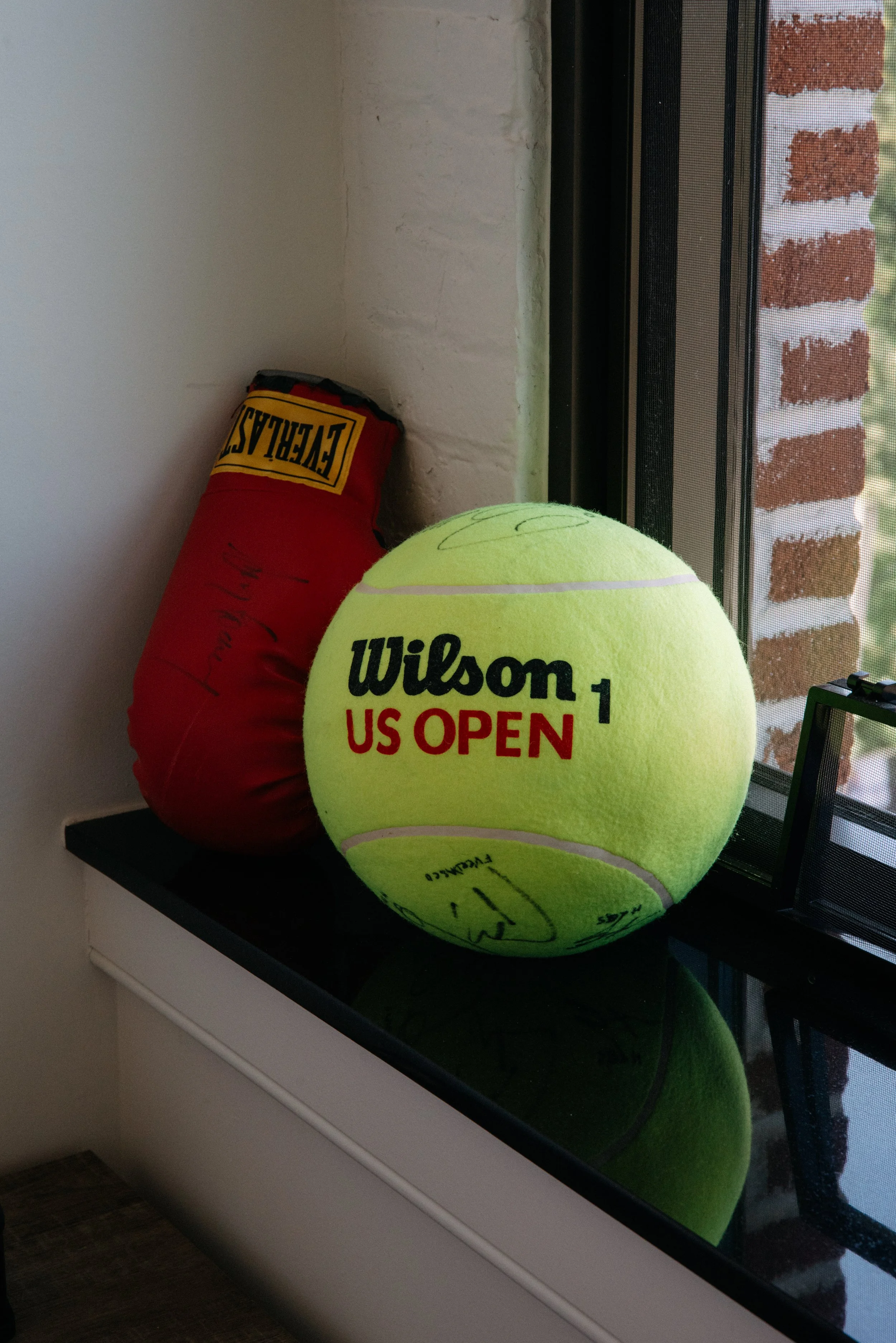A bright green Wilson tennis ball and a red Everlast boxing glove are placed on a windowsill with a brick building visible outside.