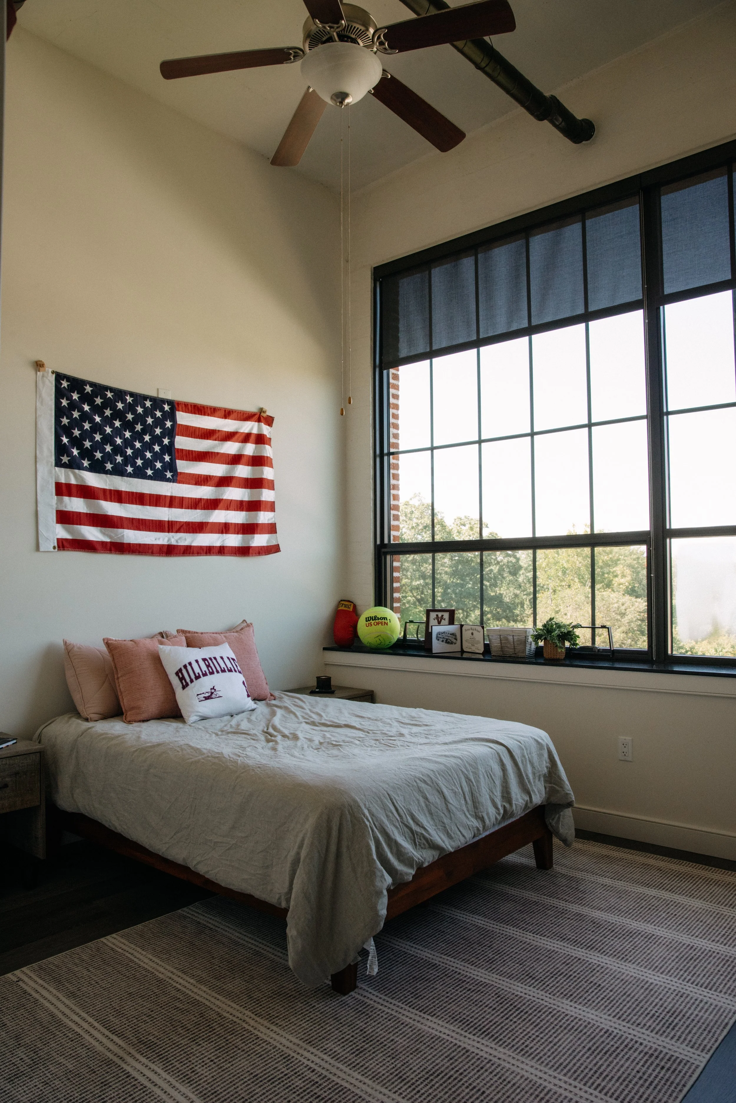 Bedroom with bed, American flag on wall, tennis ball, basketball, framed photos, small plant, nightstand, large window, ceiling fan.