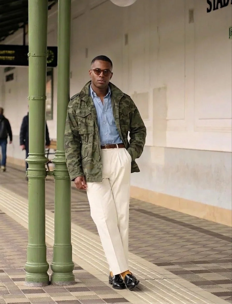 A man standing outside in front of a light blue house with a white window, surrounded by colorful plants and flowers, wearing a brown plaid coat, black turtleneck, brown pants, black loafers, and a multicolored striped scarf.