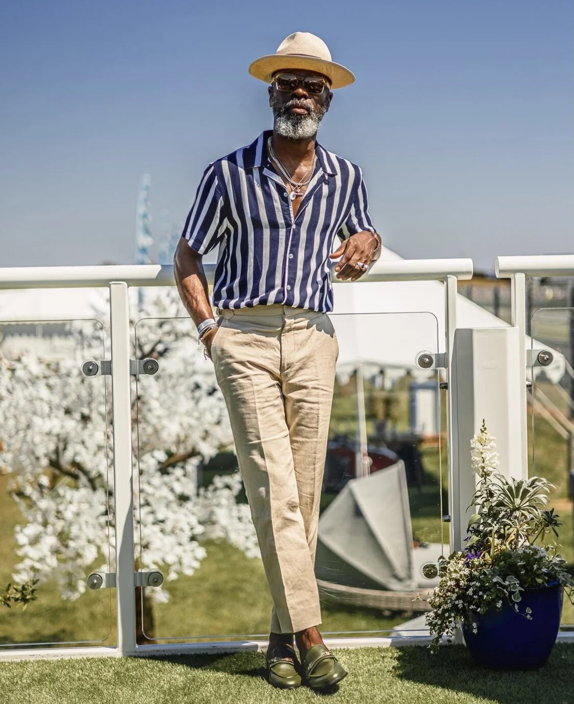 Stylish man wearing a straw hat, sunglasses, striped shirt, beige pants, and loafers, standing outdoors beside a glass railing with a basket of flowers nearby.