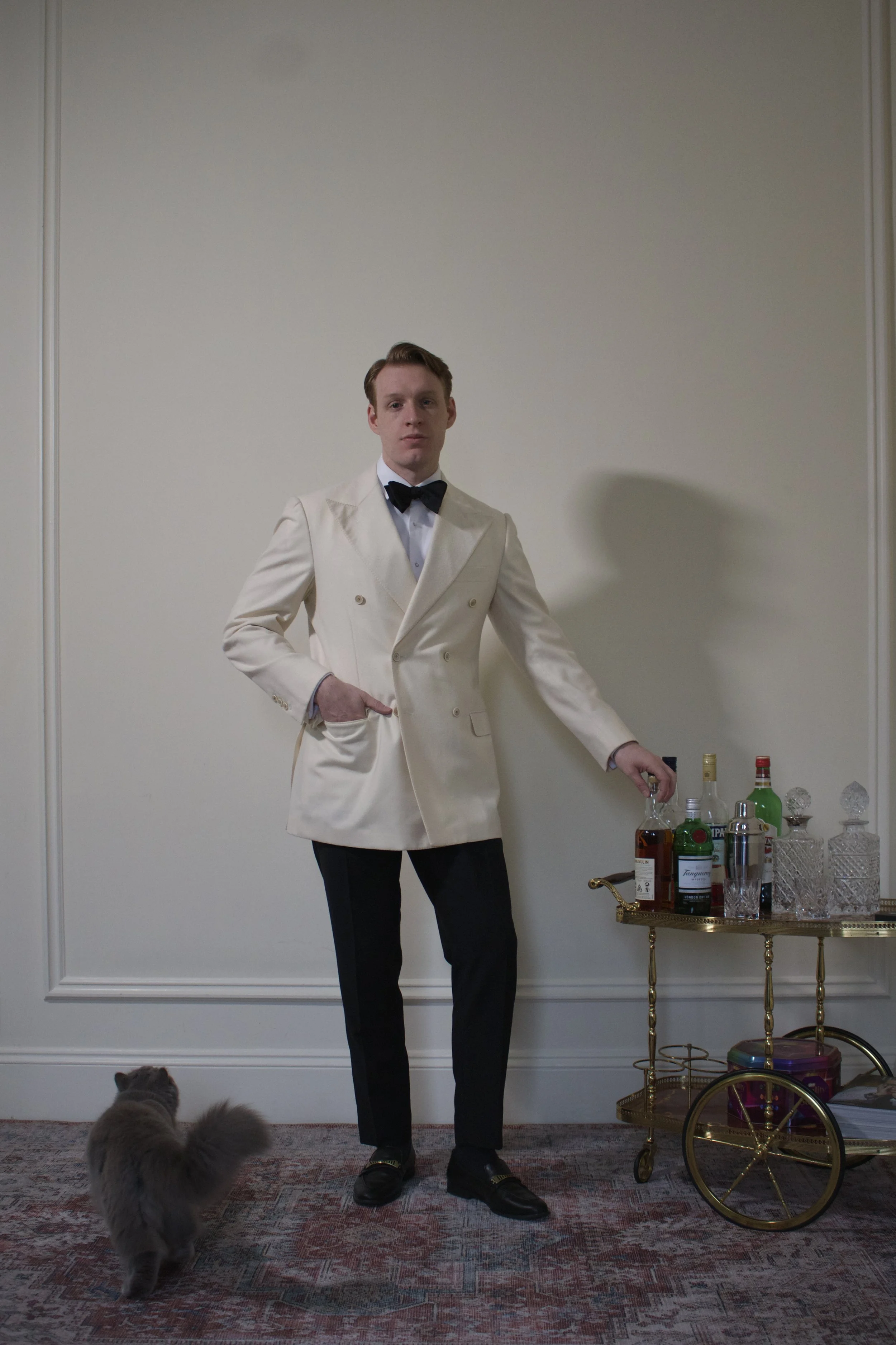 Man in a white tuxedo and black bow tie standing next to a bar cart with various liquor bottles and glassware, with a gray cat nearby, in an elegant room with a white wall.