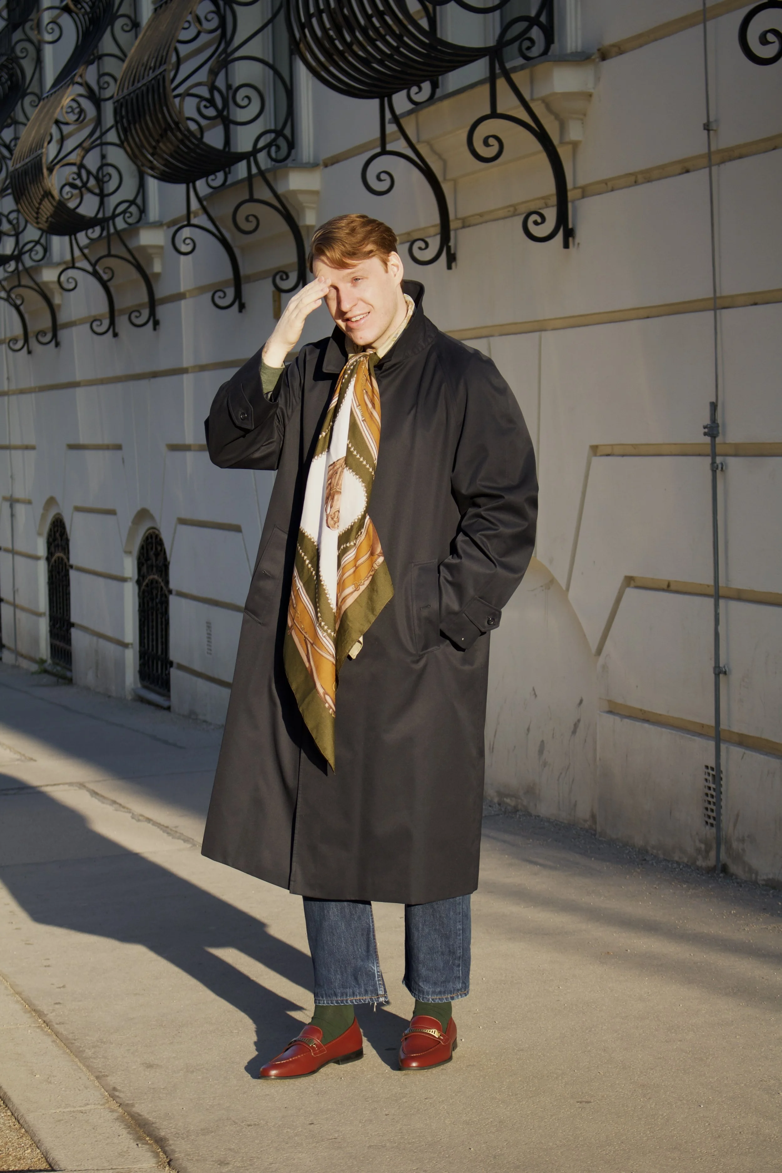 Man standing on sidewalk wearing a black trench coat, green socks, red loafers, and a patterned scarf, with hand on forehead and smiling, in front of a white building with ornamental black ironwork.