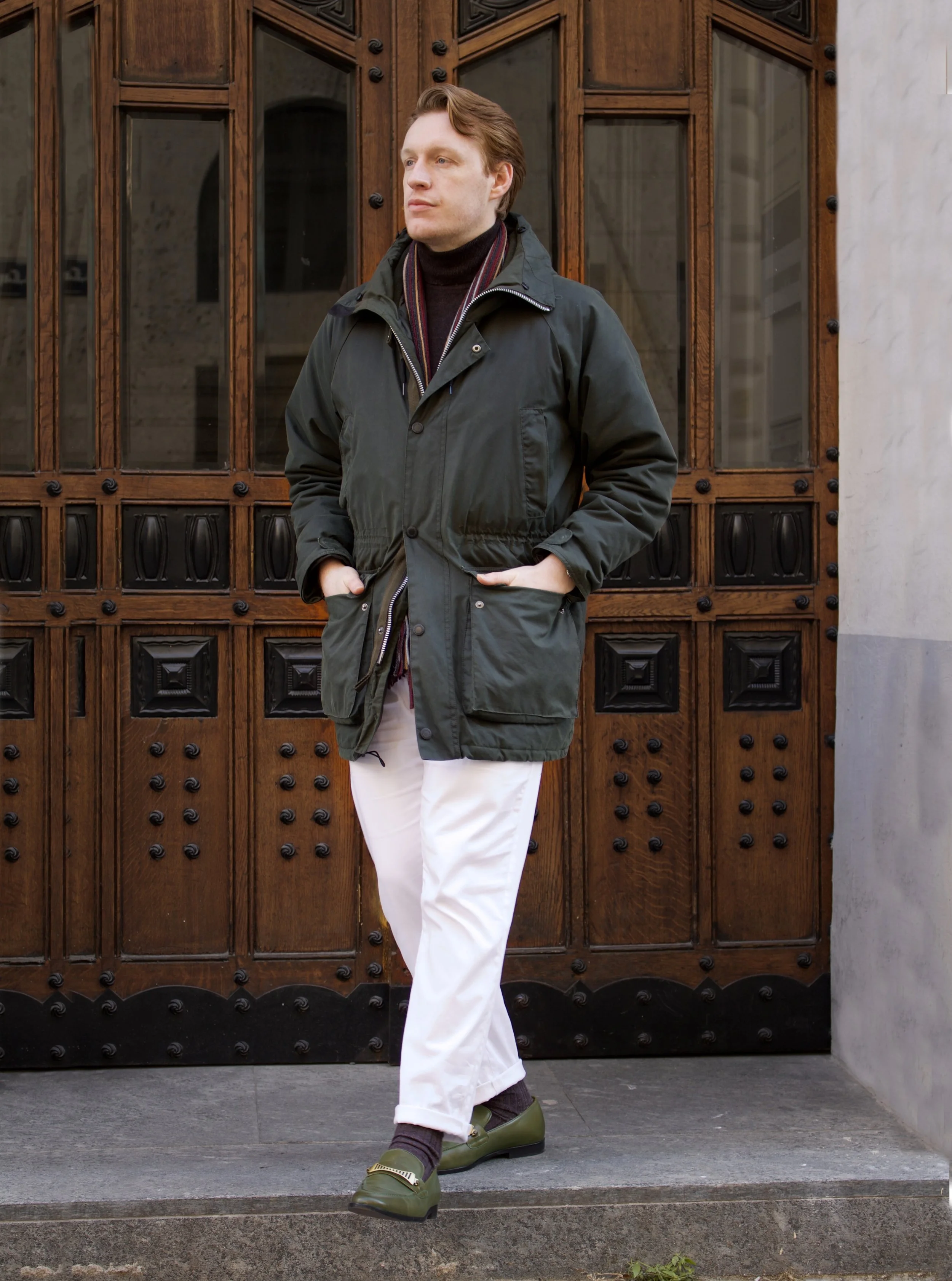 A man standing in front of a vintage wooden door, wearing a green jacket, black turtleneck, white pants, and olive green shoes.