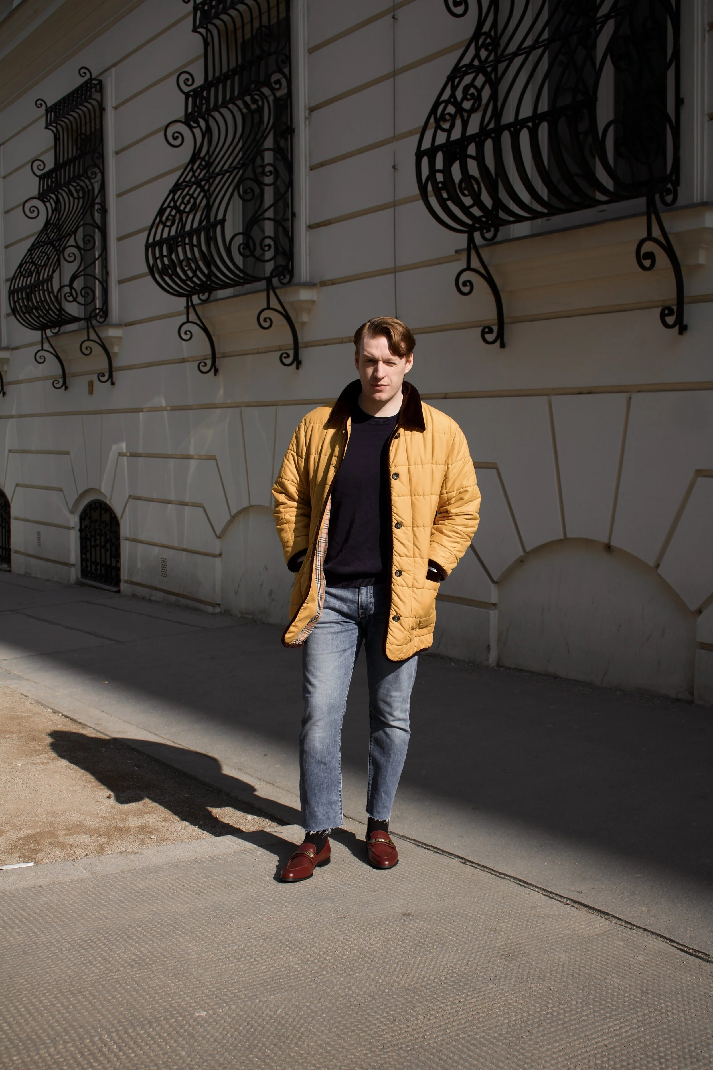 Young man wearing a mustard yellow jacket, black shirt, and jeans walking on the sidewalk in front of a white building with decorative black window grilles.