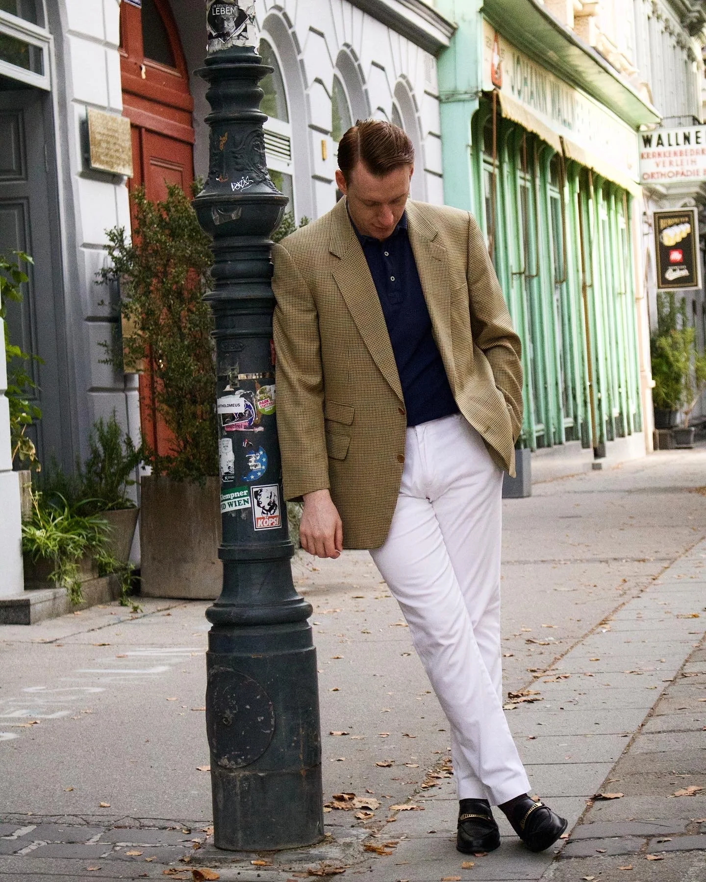 A man in a beige jacket, dark shirt, and white pants leaning against a black street lamp on a sidewalk in front of colorful storefronts.