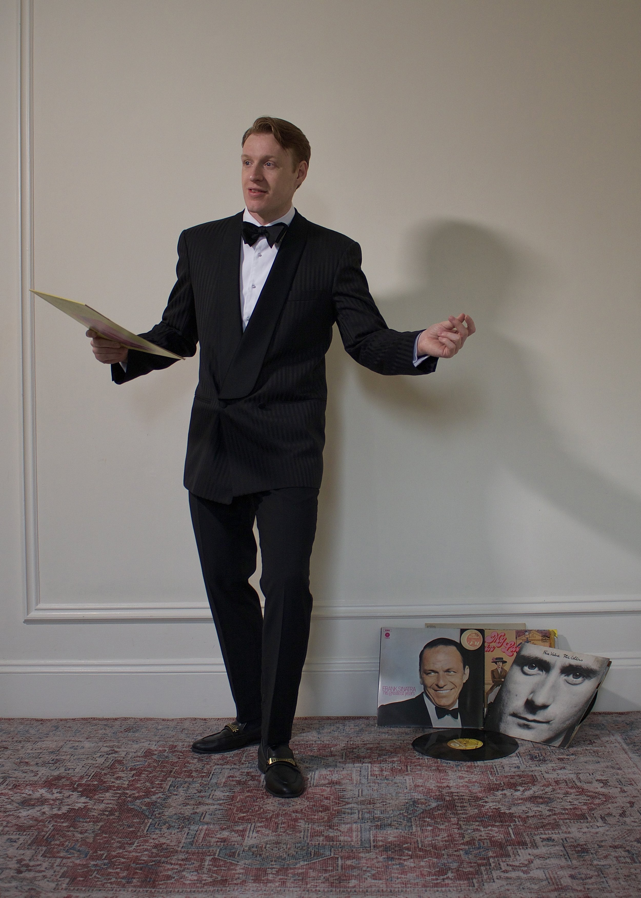 A man in a tuxedo standing against a white wall holding a booklet or menu. There are vinyl records on the floor near him, including one with a portrait of Frank Sinatra.