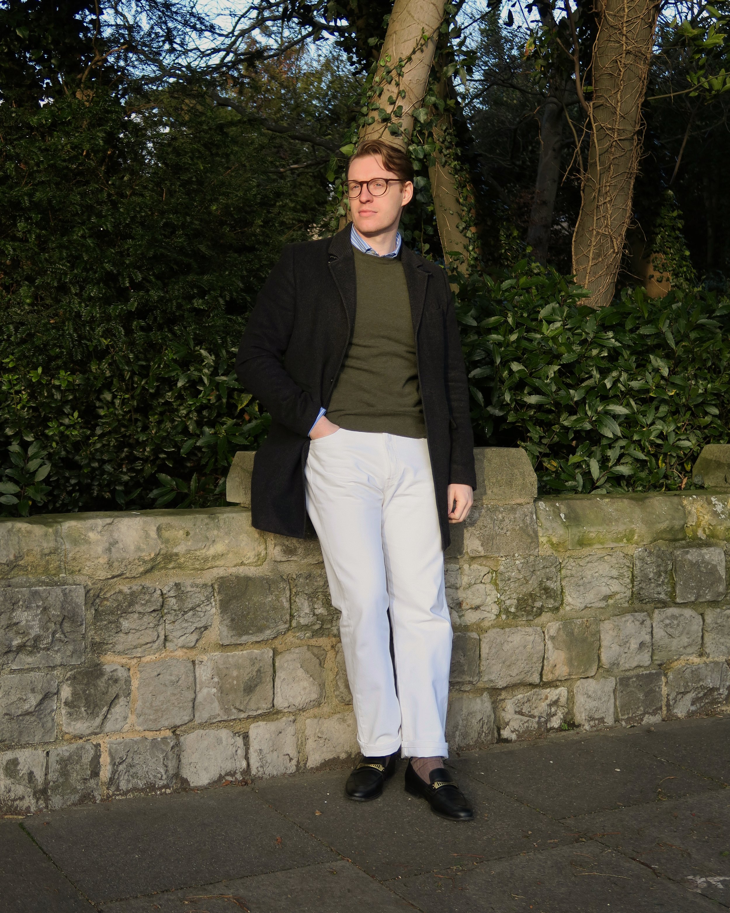 A young man wearing glasses, a dark coat, green sweater, striped shirt, and white pants standing outdoors near a stone wall with green foliage and trees in the background.