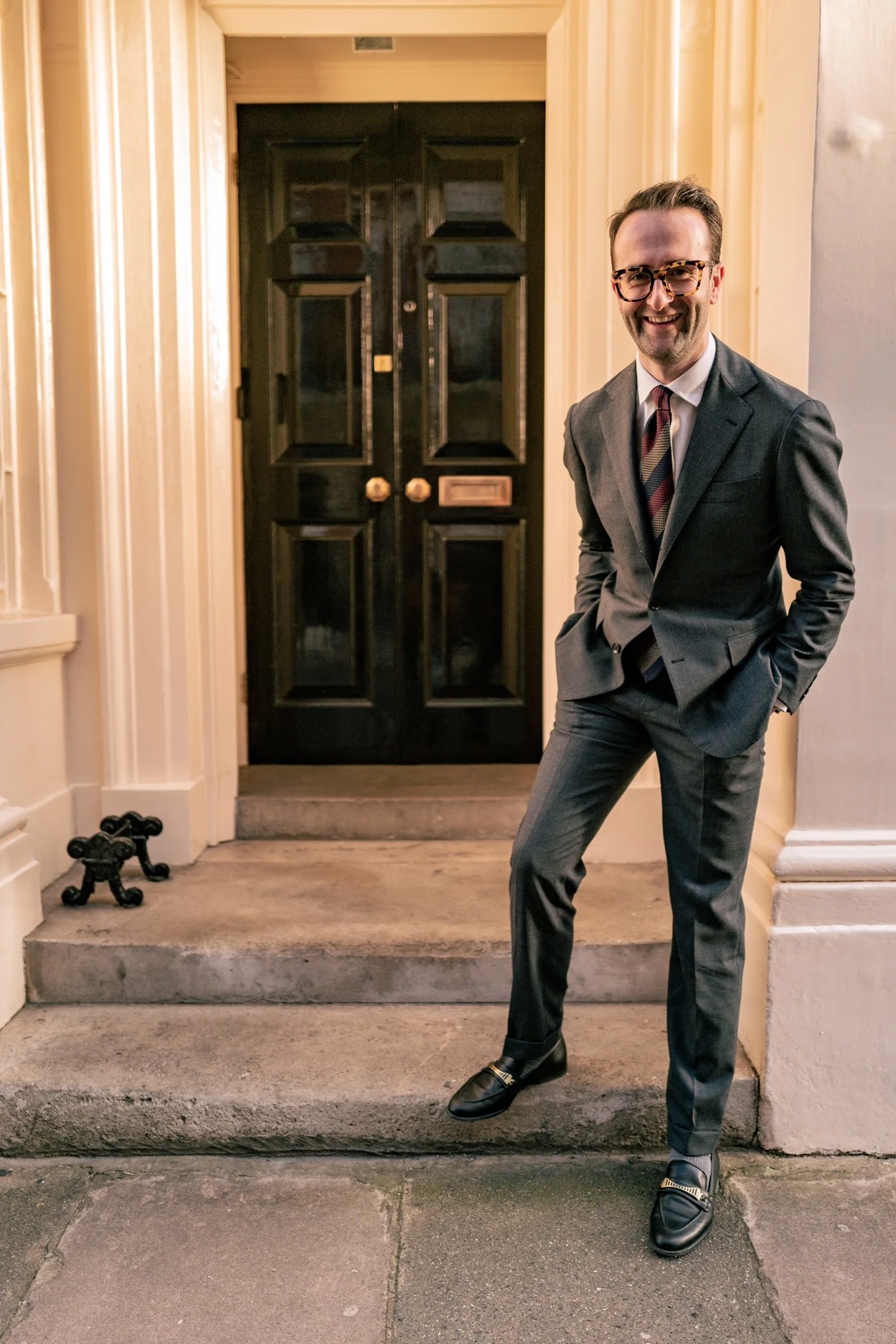 A man wearing glasses and a dark gray suit with a striped tie standing on the steps outside a black door with gold knobs and mail slot.