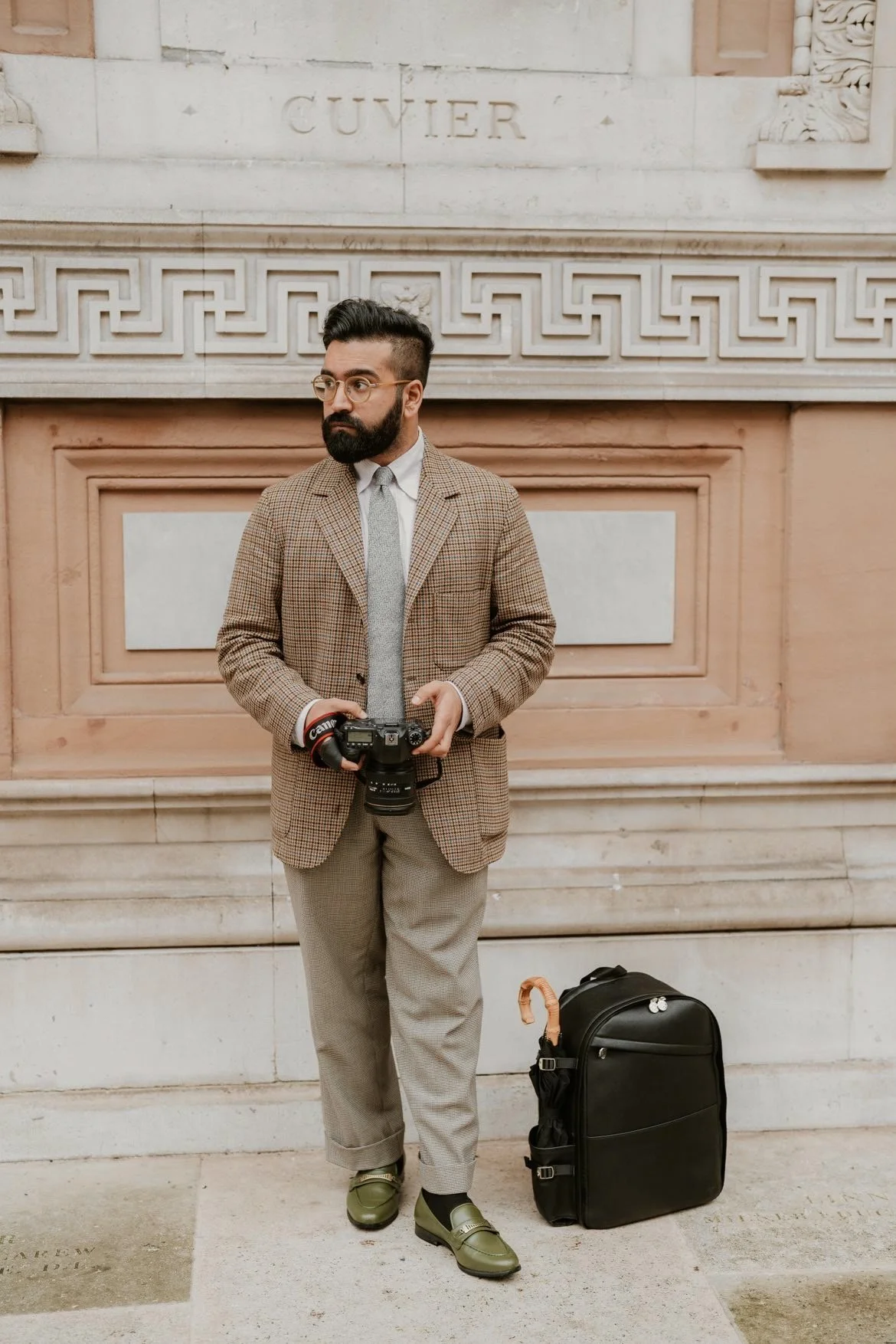 A man dressed in a checked blazer and gray trousers holding a Canon camera stands in front of a stone wall with intricate designs and the word 'Cuvier' engraved above.