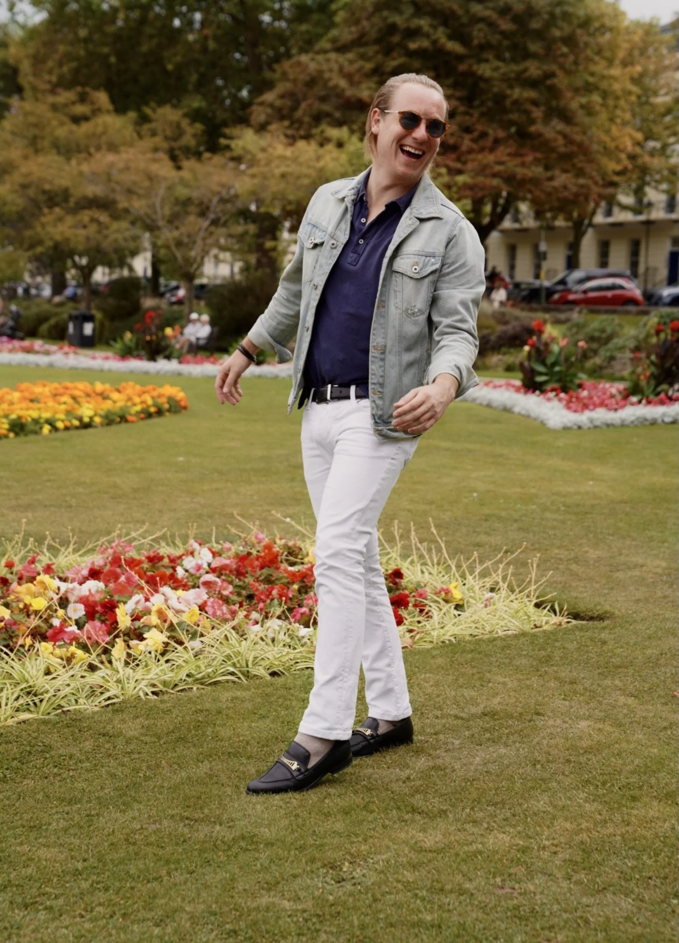 A man with long hair and sunglasses smiling and laughing while standing on a lush green lawn in a park with colorful flower beds and trees in the background.