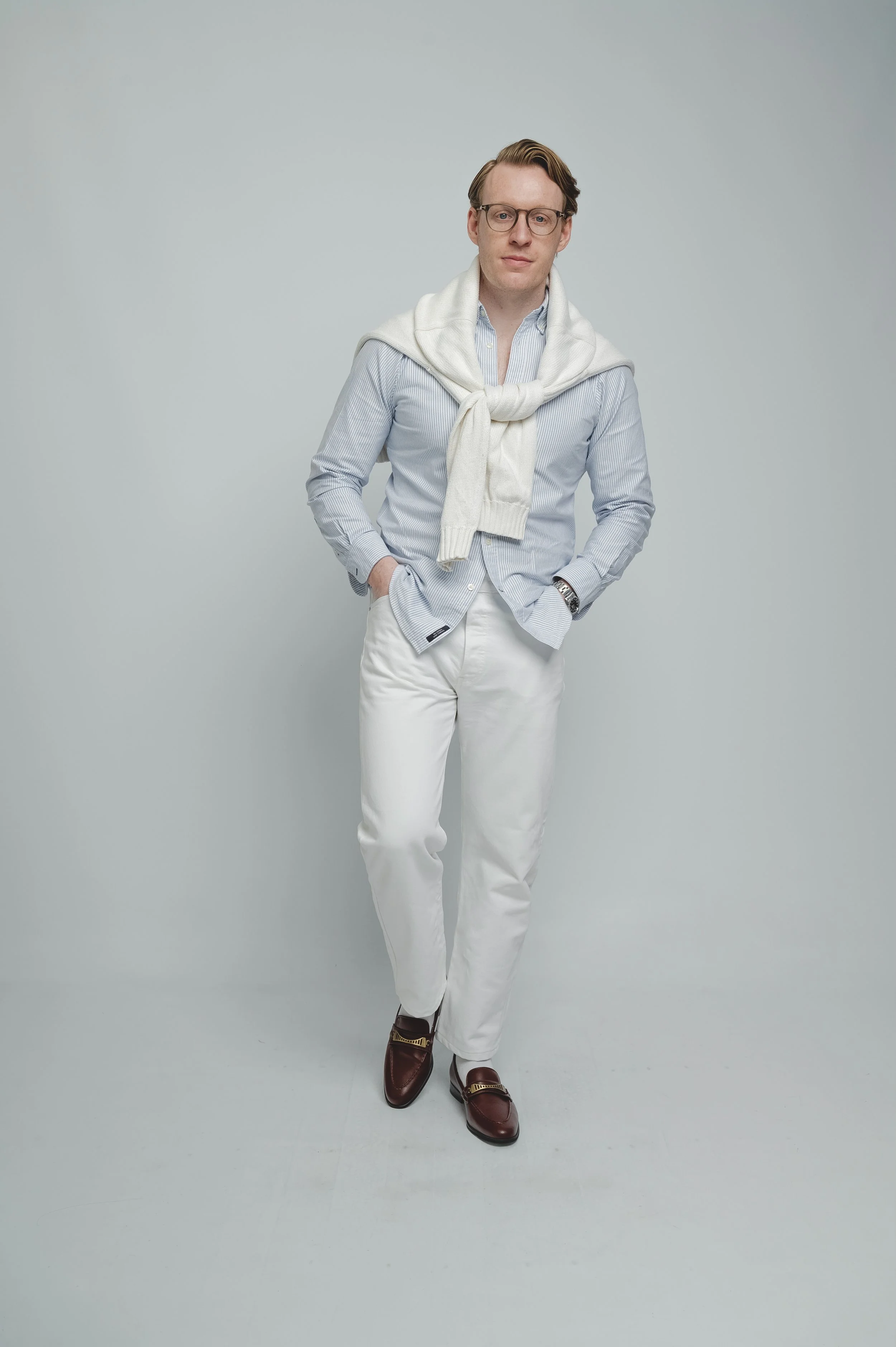 A young man with glasses and styled hair standing with hands in pockets against a plain light gray background, wearing a light blue striped button-up shirt, cream sweater draped over shoulders, white pants, and brown loafers.