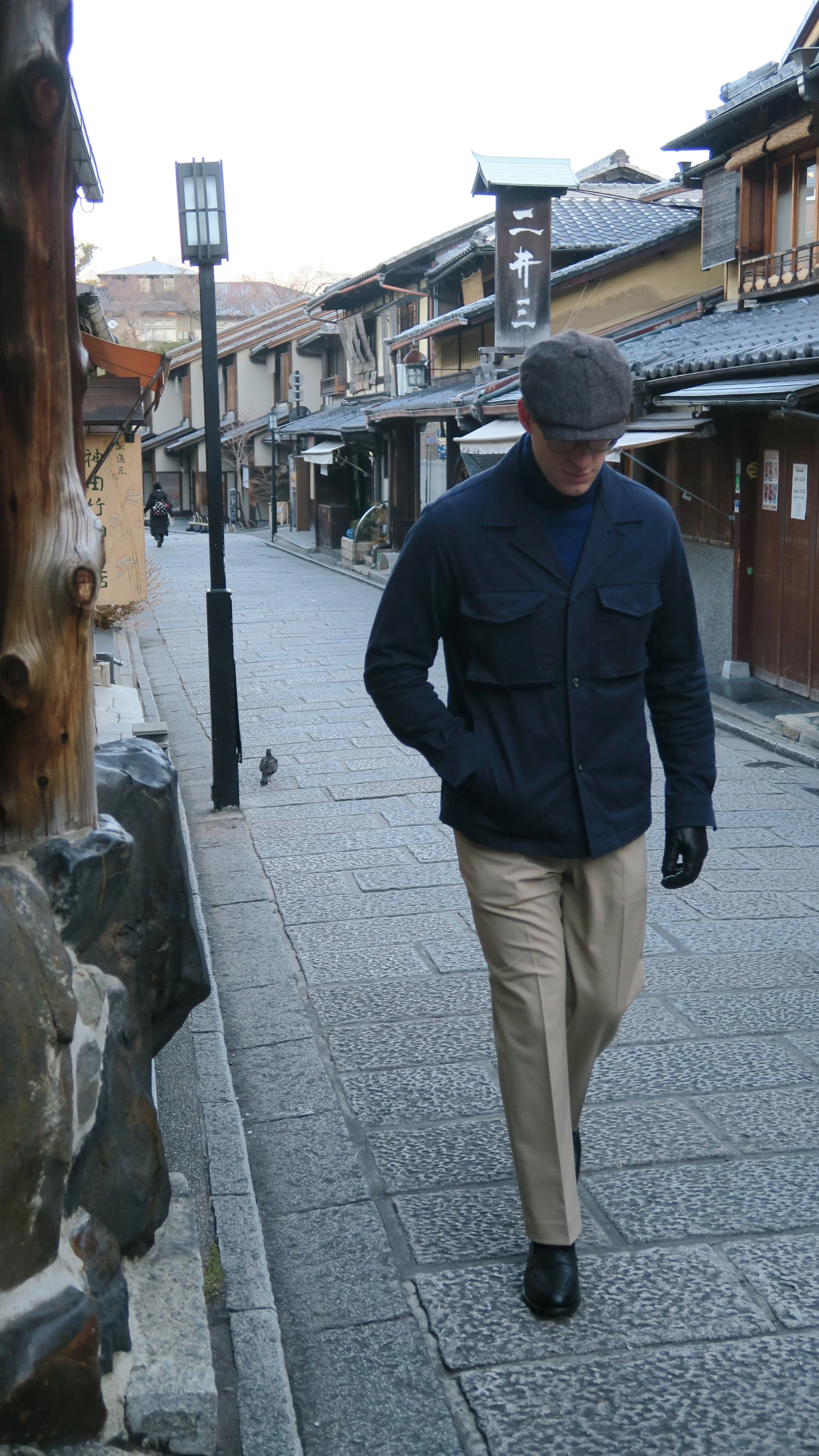 A man in a gray cap, dark jacket, jeans, and gloves walks along a traditional Japanese street with wooden buildings and tiled roofs.