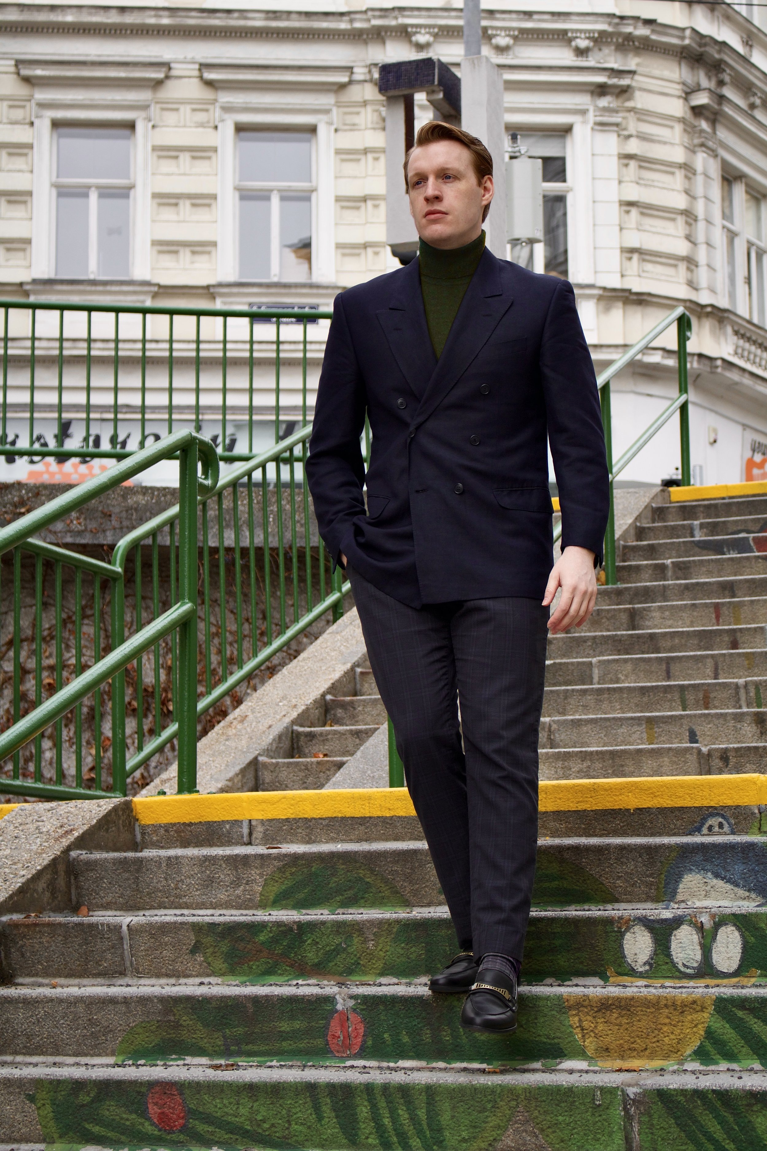 A young man with short brown hair walking down painted concrete stairs outdoors, wearing a dark navy blazer, dark checkered pants, and black shoes. In the background are old buildings with large windows and green railings.