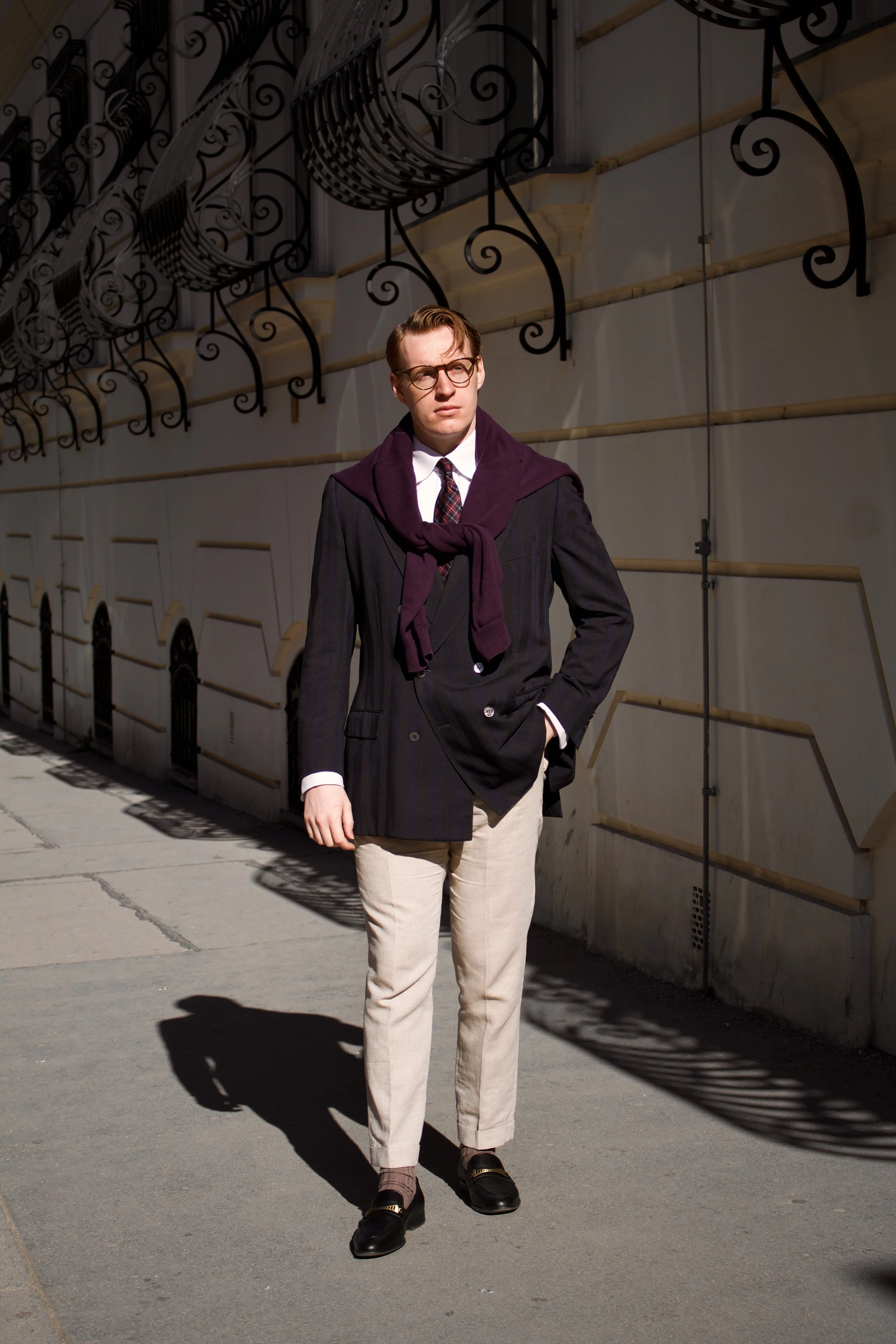 Young man with glasses wearing a dark blazer, beige pants, and loafers stands on a sidewalk in sunlight, with a wall and ornate metal design overhead casting shadows.
