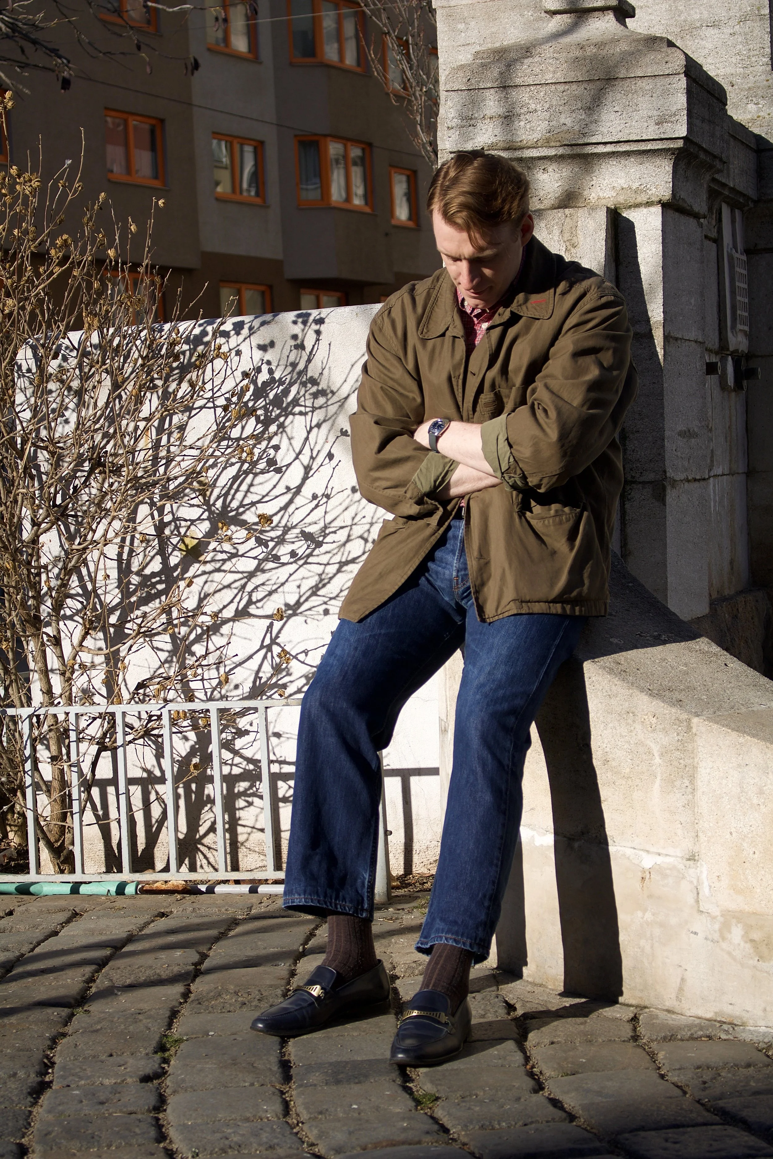 A young man with brown hair stands on a cobblestone street, leaning against a stone structure outdoors, wearing a brown jacket, blue jeans, and black loafers. He has his arms crossed and appears to be looking down, with shadows cast by nearby trees o