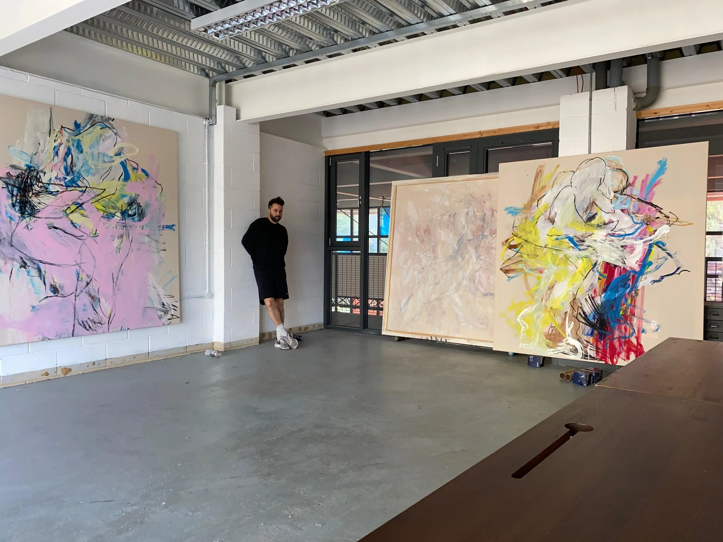 An art studio with abstract paintings on display and a man leaning against a white brick wall.