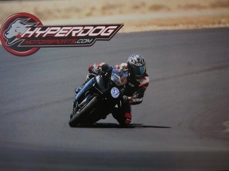 A motorcycle racer in full gear riding a black motorcycle on a racetrack, with a Hyperdog Motorsports.com logo in the upper left corner.