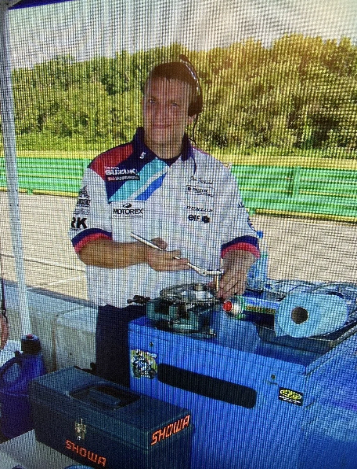A man in a racing team uniform with various sponsor logos, wearing a headset, working with a device at a table outdoors with green trees and a fence in the background.