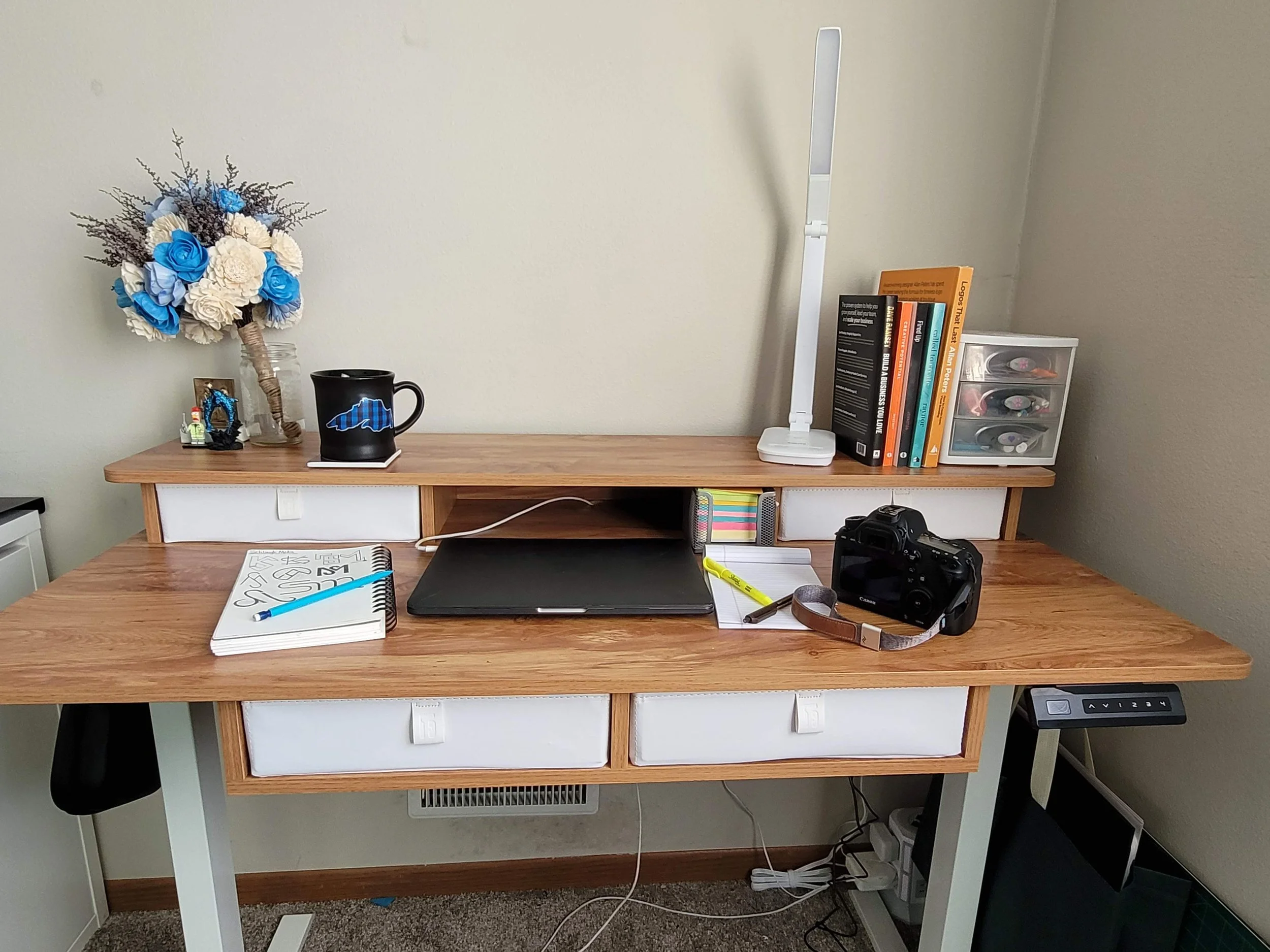 A wooden desk with white drawers, holding a black mug with a blue graphic, a closed laptop, a notebook with a blue pen, a camera, and various office supplies. A tall white lamp and a small plastic storage container with multicolored drawers are on the desk. A floral arrangement with blue and white flowers in a glass vase is on the left, and a row of books with a white digital clock is on the right.
