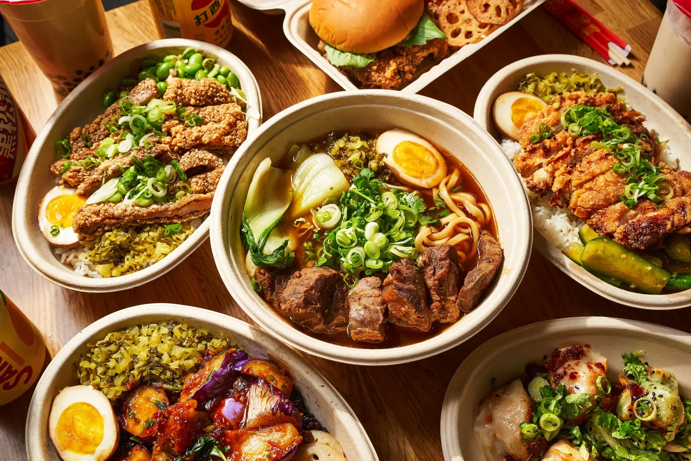 A variety of Asian dishes including bowls of ramen with boiled eggs and green onions, fried chicken with lettuce and lotus root, and other small dishes on a wooden table.