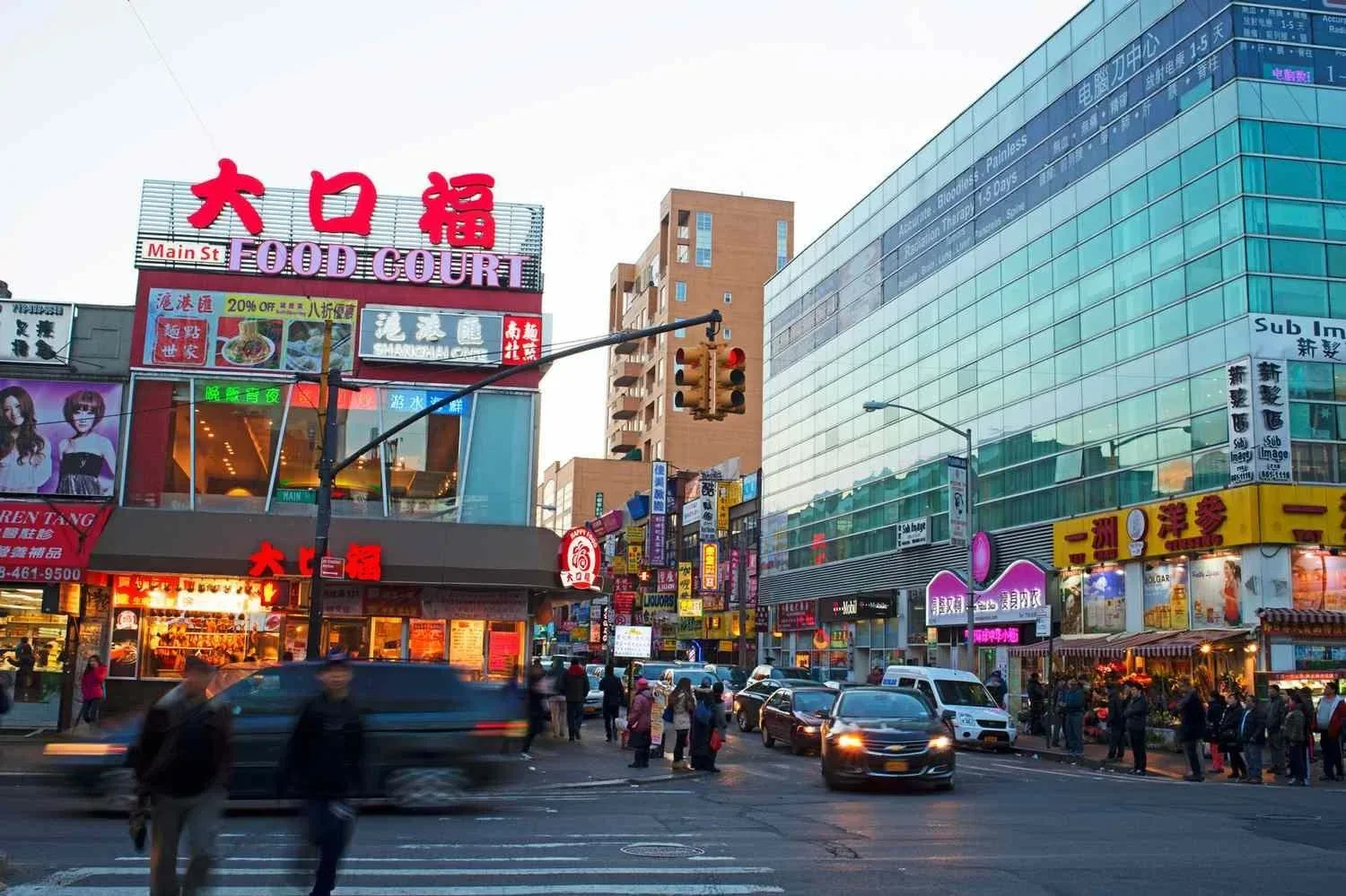 A busy city street scene at dusk with shops, colorful signs, and people crossing the street. There are cars and pedestrians, with a large building featuring glass windows on the right and smaller buildings with bright signage on the left.