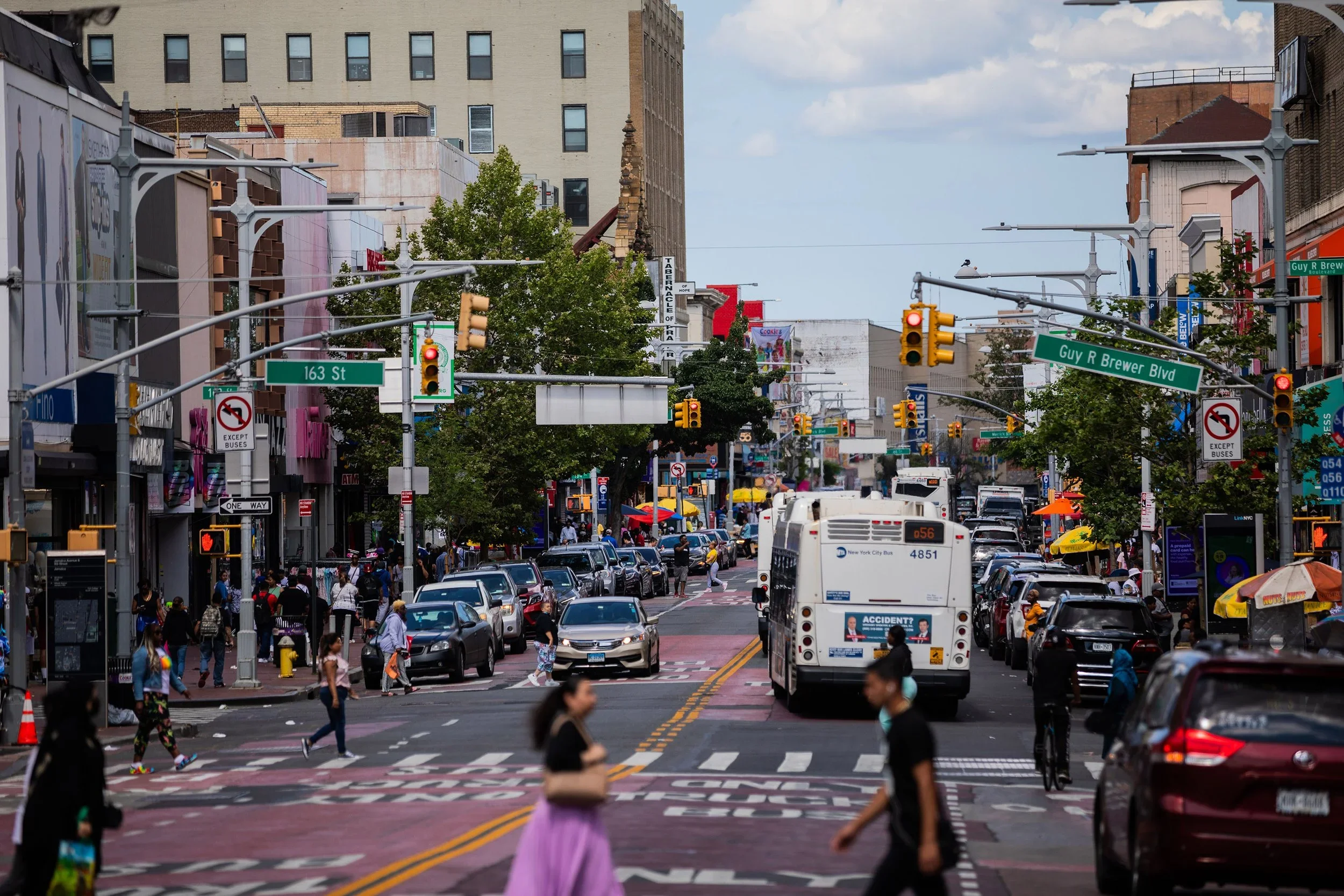 A busy city street filled with cars, buses, and pedestrians. Street signs indicate 163rd Street and Guy R. Brewer Boulevard. Pedestrians cross at the crosswalk, some carrying shopping bags. Buildings and advertisements line the street, with traffic lights and street lamps hanging overhead.
