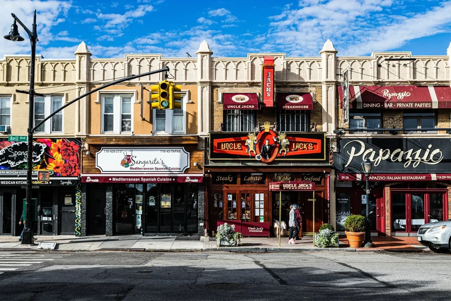 City street view with storefronts for Uncle Jack's steakhouse, Sangarita's Mediterranean Spanish Cuisine, and Papazzio Italian Restaurant, with pedestrians and traffic lights under a partly cloudy sky.