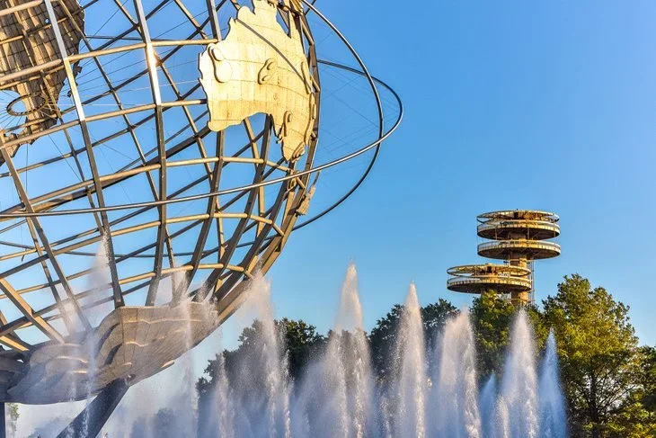 The image shows the Space Needle in Seattle, Washington, with the iconic Globe at the top and a nearby fountain with water jets, and trees against a clear blue sky.