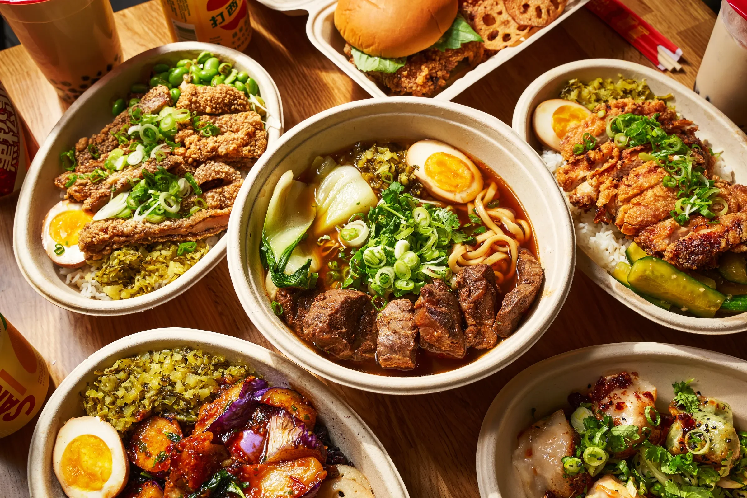 A variety of Asian-inspired dishes in individual bowls, including ramen with beef, eggs, and greens, fried chicken with rice, beef with green vegetables, and curry with eggplant, served on a wooden table with drinks and condiments.