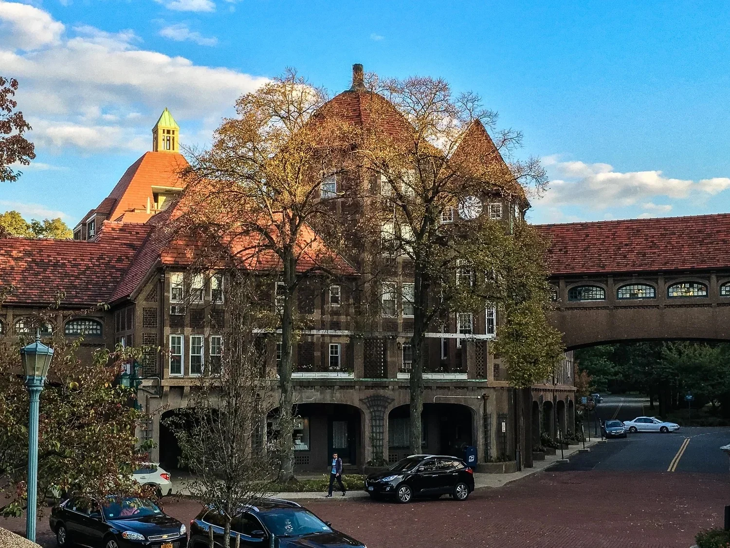 A historic brick building with a tower, trees partially covering it, cars parked in front, and a bridge connecting to another part of the building. A person walks on the sidewalk beneath the trees.
