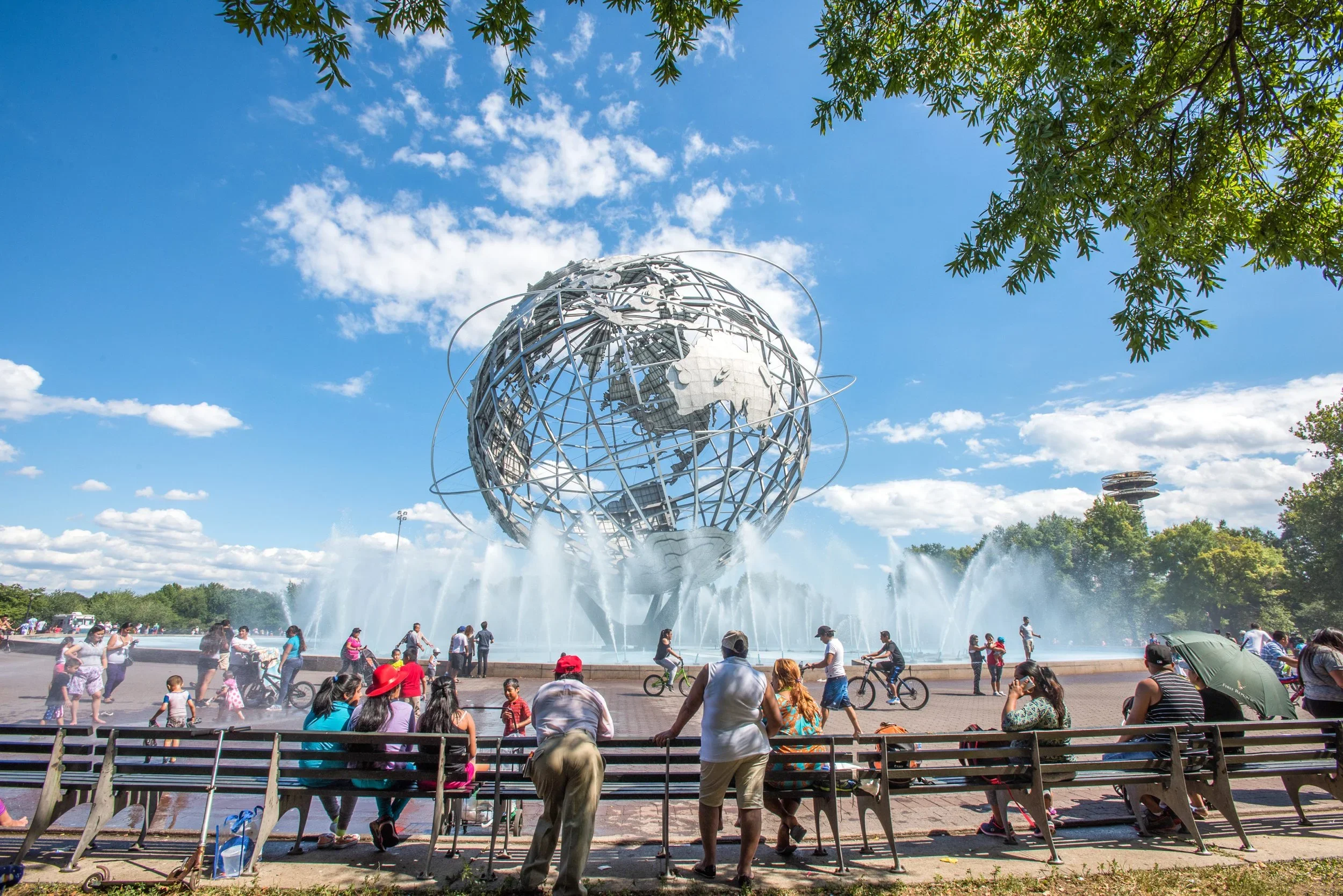 The image shows a large crowd of people gathered around the fountain at the Unisphere, a giant spherical steel representation of Earth, with trees and a partly cloudy sky in the background.