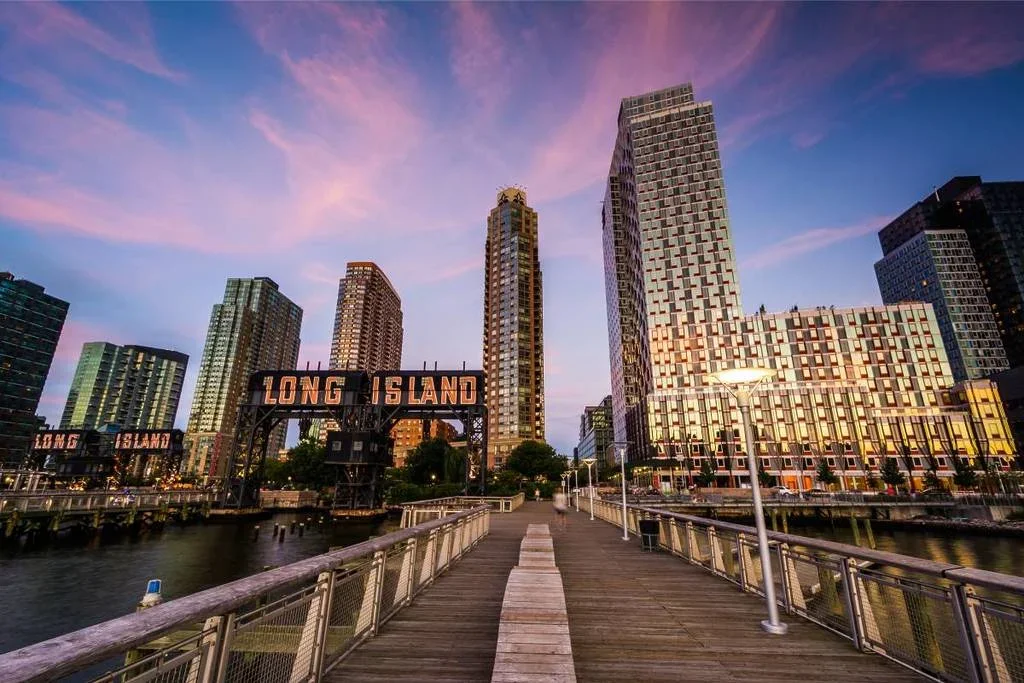 Skyline view of Manhattan buildings from a pier with a sign that says "Long Island" at sunset.