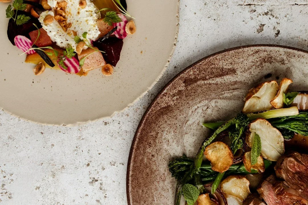 Two plates of gourmet food on a textured white surface, one with roasted vegetables and meat, the other with a colorful salad with radishes, nuts, and herbs.