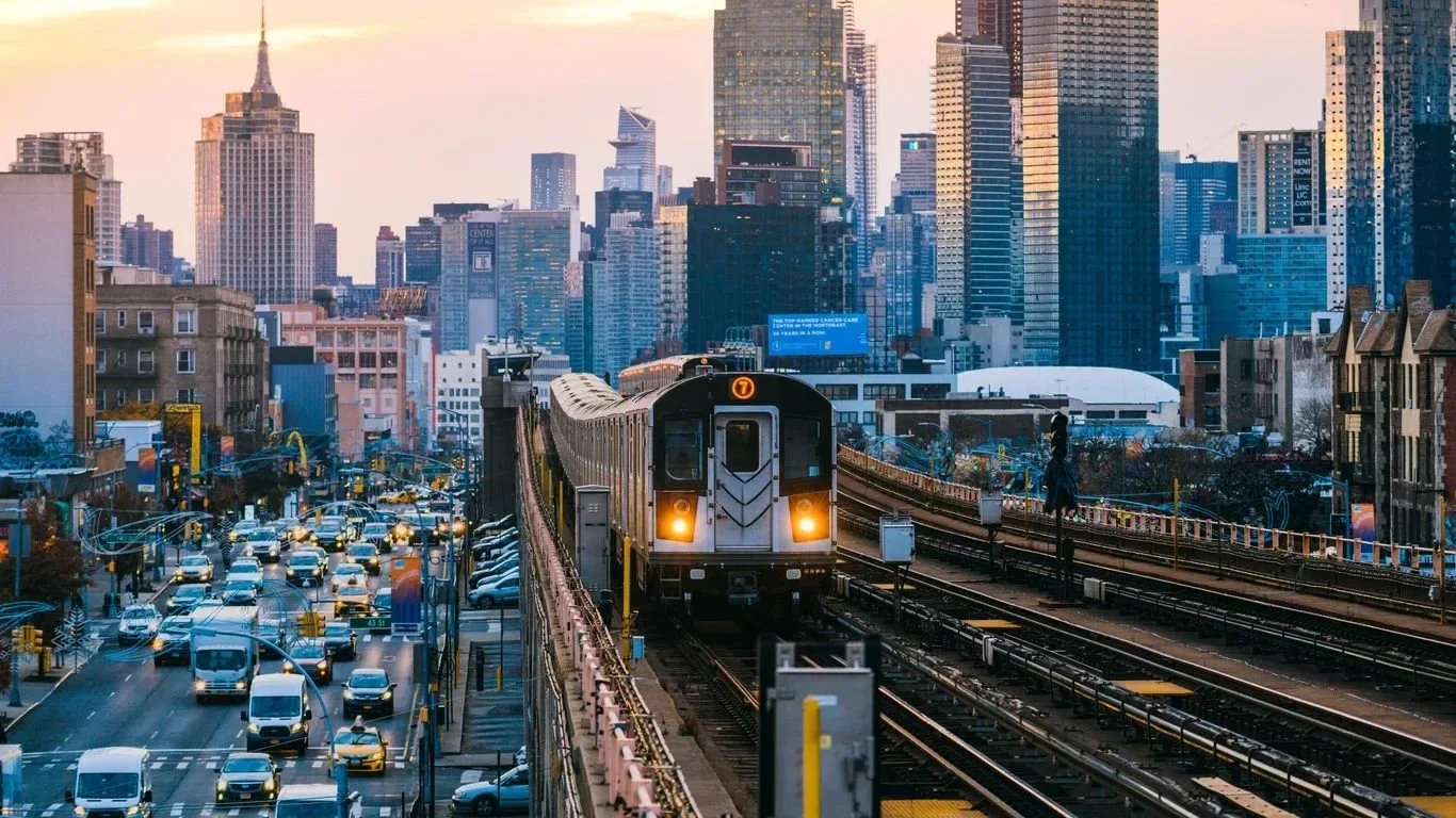 A cityscape featuring a busy street and a train on an elevated track with tall skyscrapers in the background during sunset or sunrise.