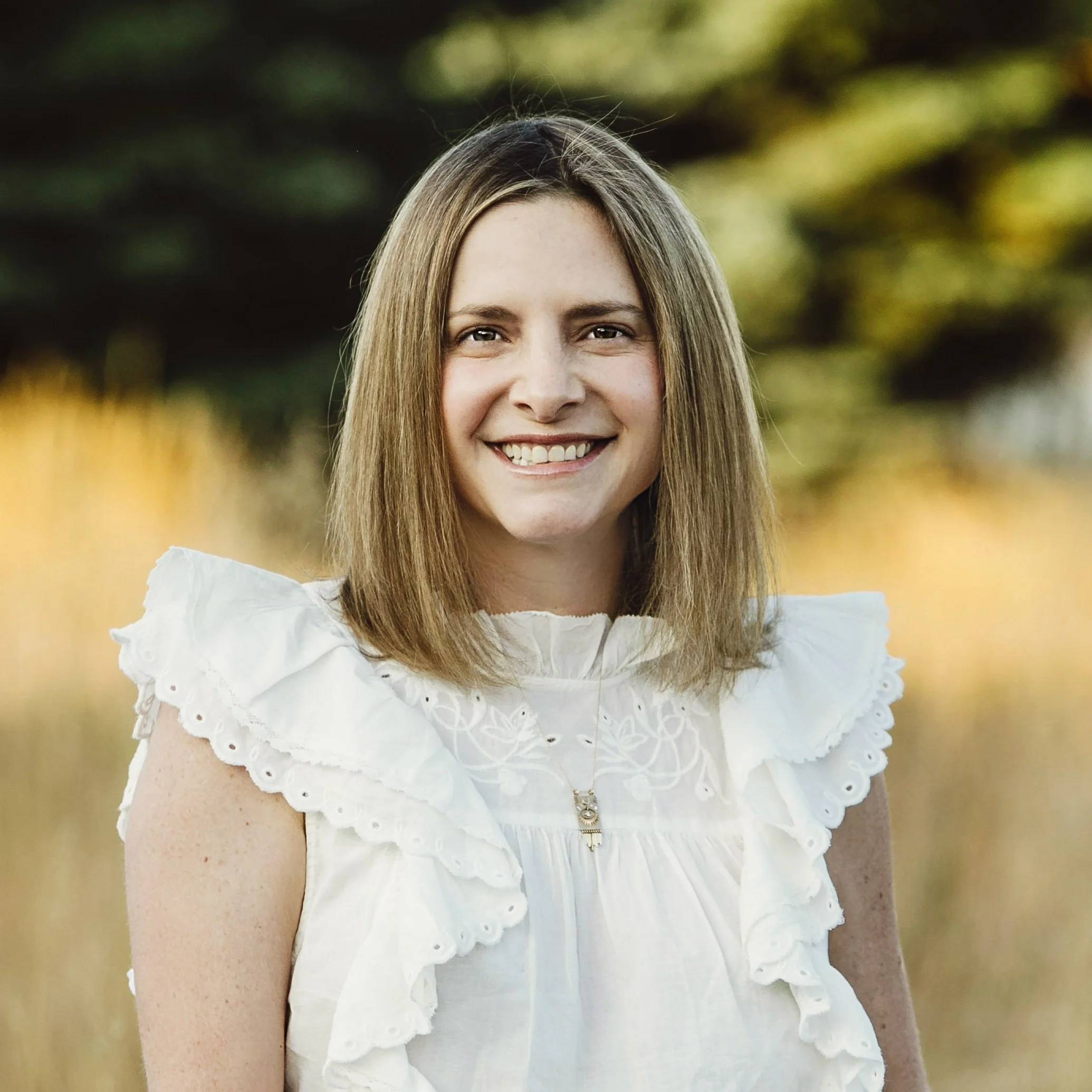 headshot of Michaela Hackner, a white woman wearing a white top standing outside. She has shoulder-length blond hair and smiles. 